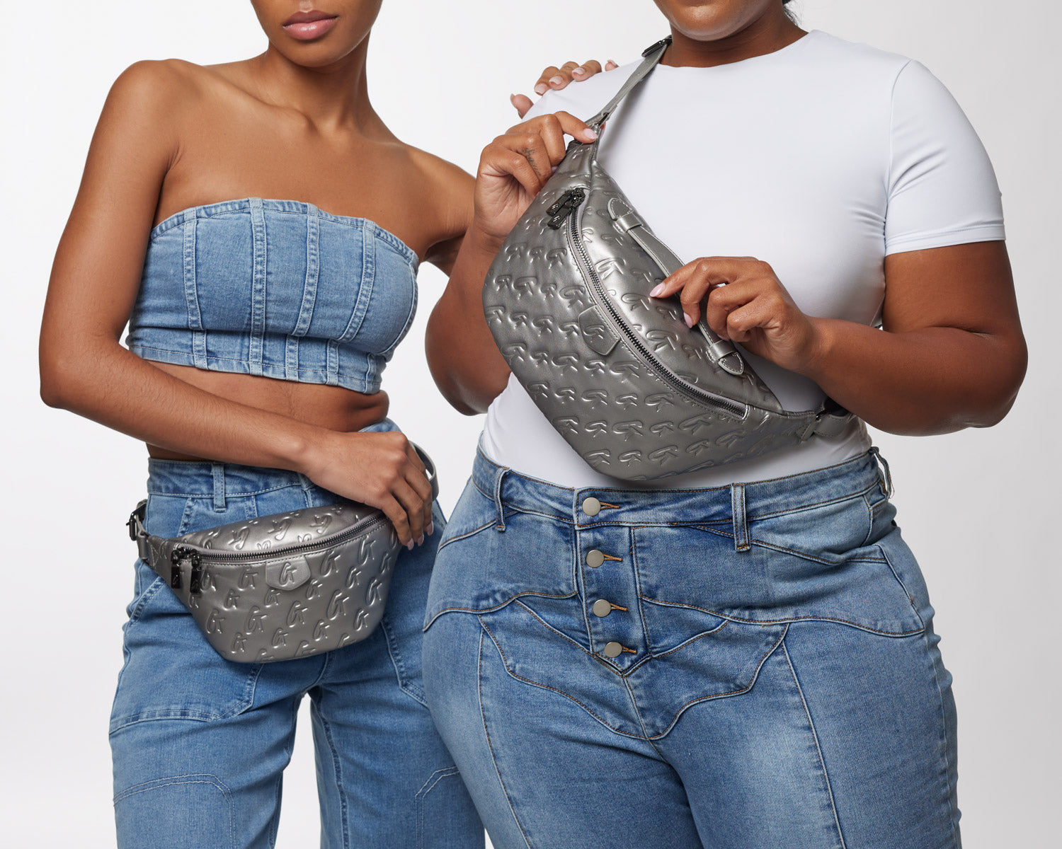 Two women display silver Monogram Fanny Packs with embossed logo patterns, styled with casual denim outfits.