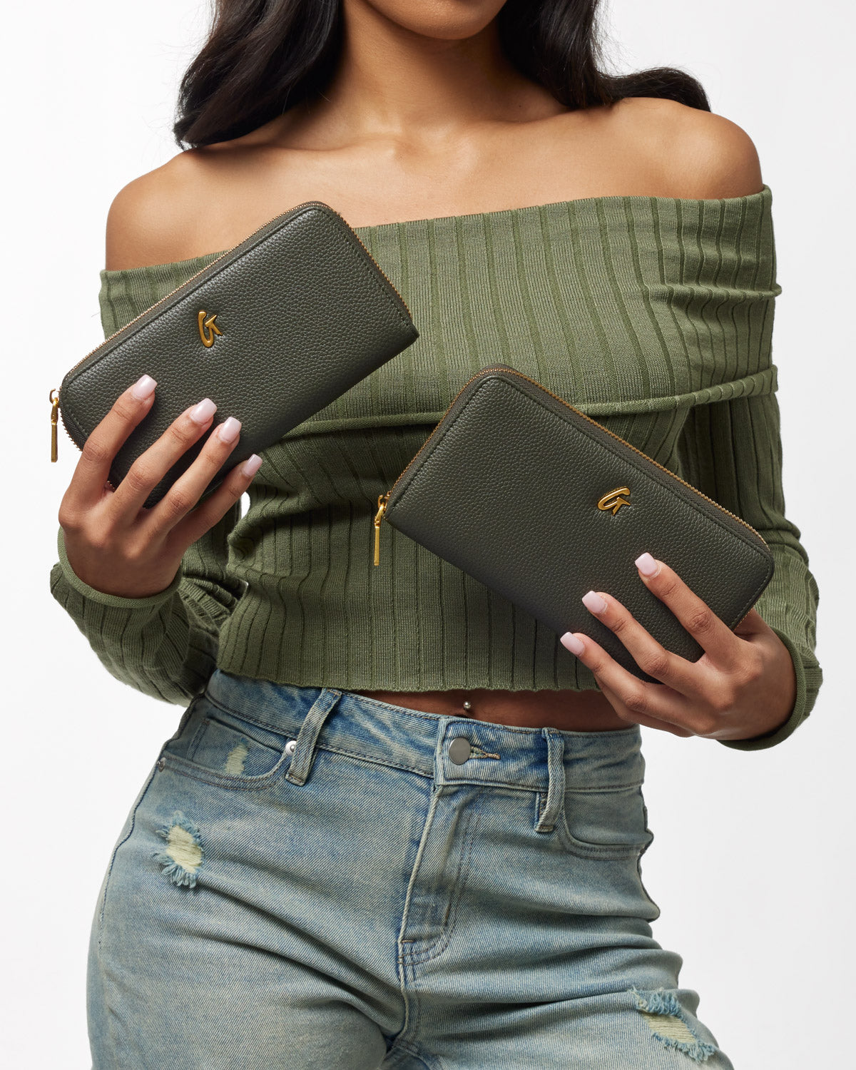 Olive green pebble leather wallets with gold zipper and logo, held by a woman in a green off-shoulder top and distressed jeans.