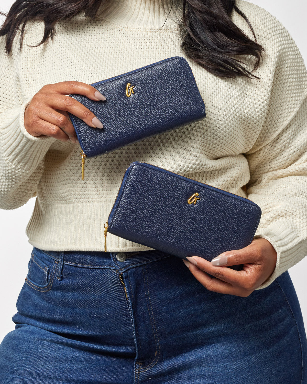 Two navy blue pebble-textured wallets with gold zippers and a gold "G" logo, held by a woman in a cream sweater and jeans.