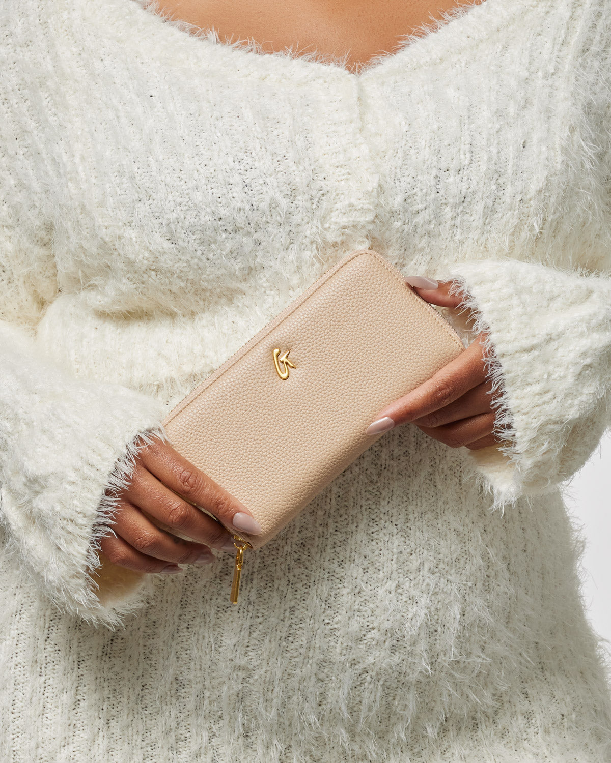 A person holding a nude-colored pebble leather wallet with a gold logo and zipper, wearing a cozy white sweater.