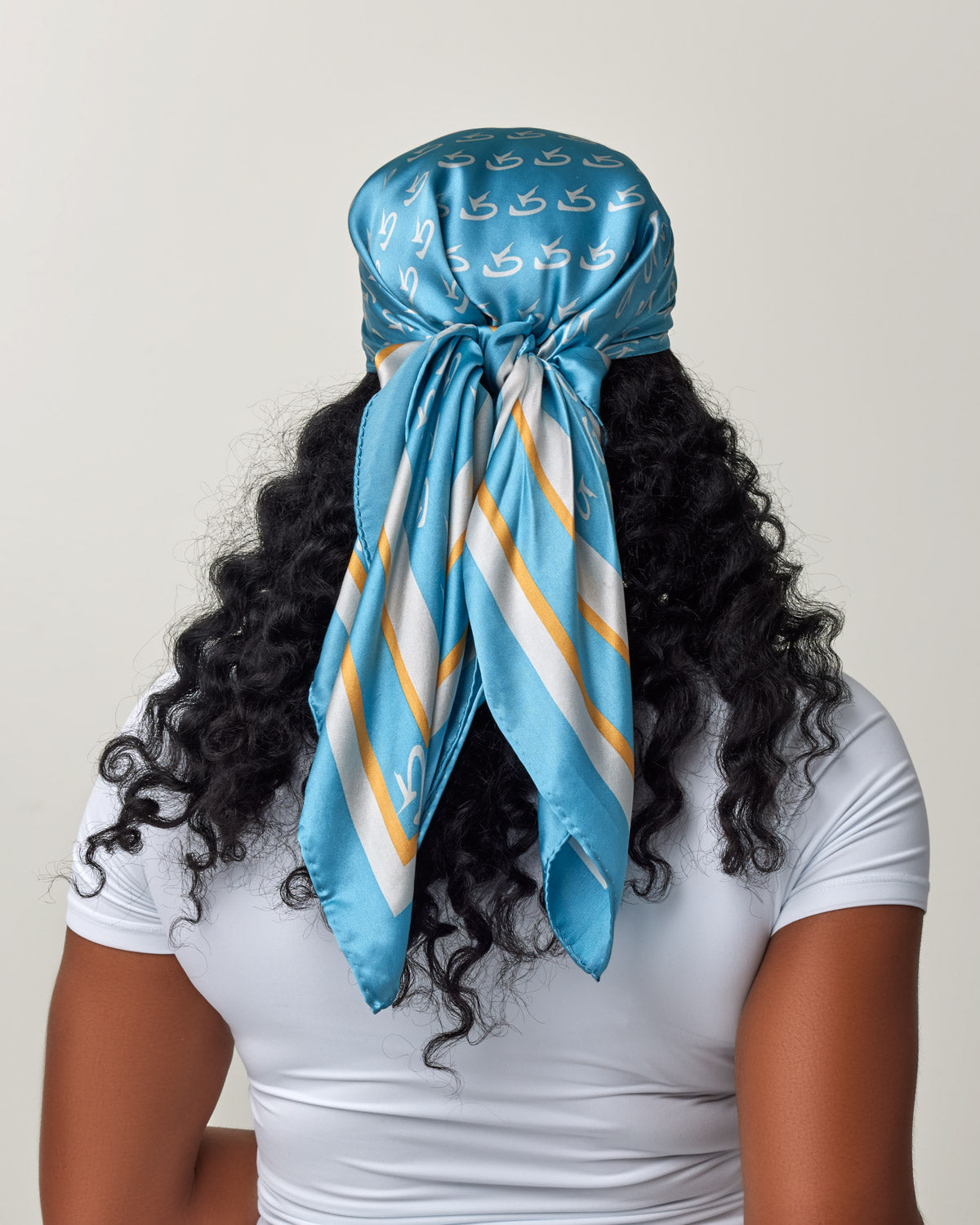 Large blue silk scarf with gold monogram pattern, tied at the back, worn over curly hair and a white top.