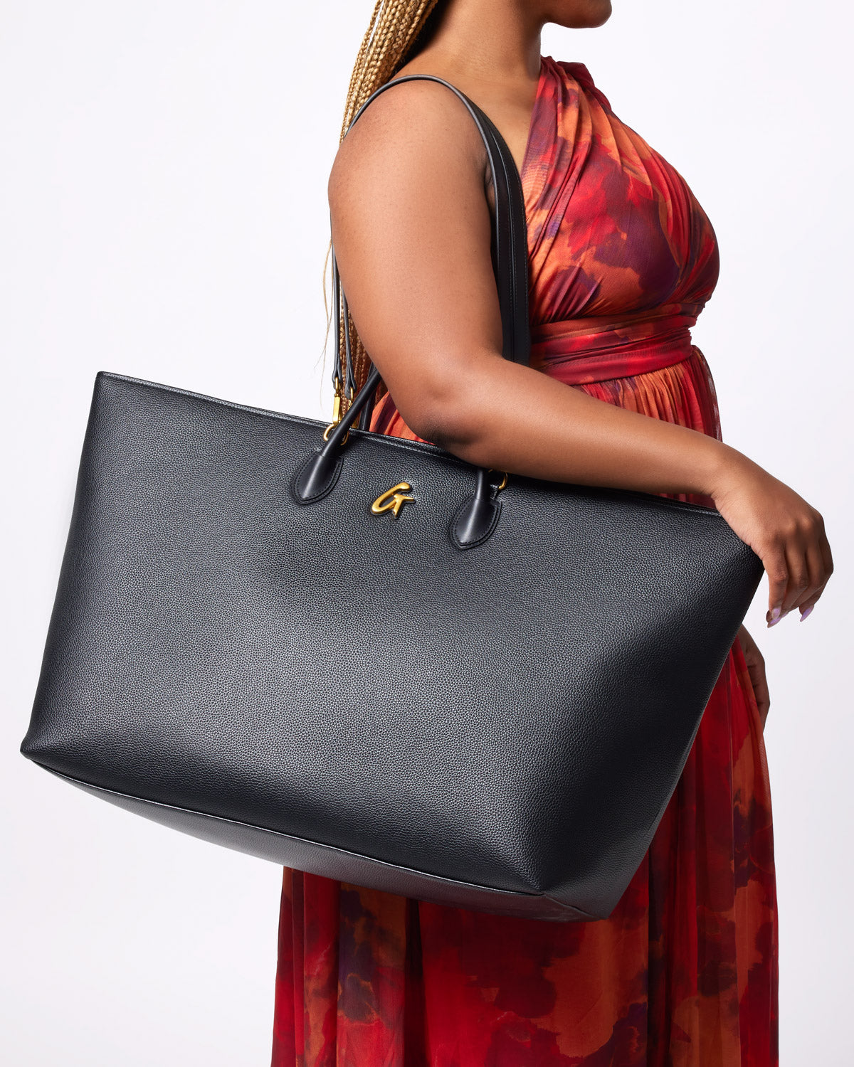 Black jumbo pebble tote bag with gold logo, large size, and dual top handles, carried by a woman in a red patterned dress.