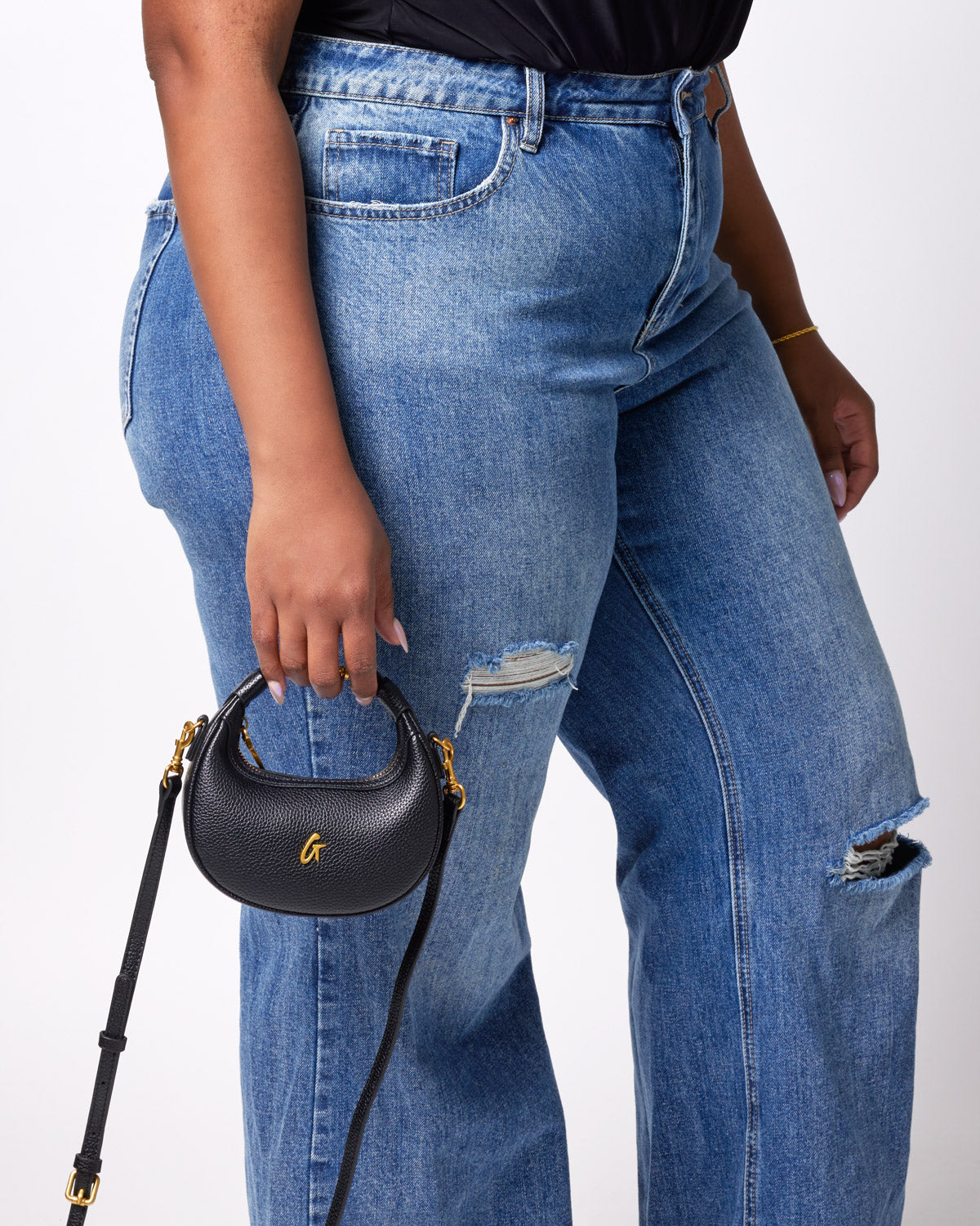 Black Nano Pebble Hobo Bag with gold hardware, held by a person wearing ripped blue jeans and a black top.