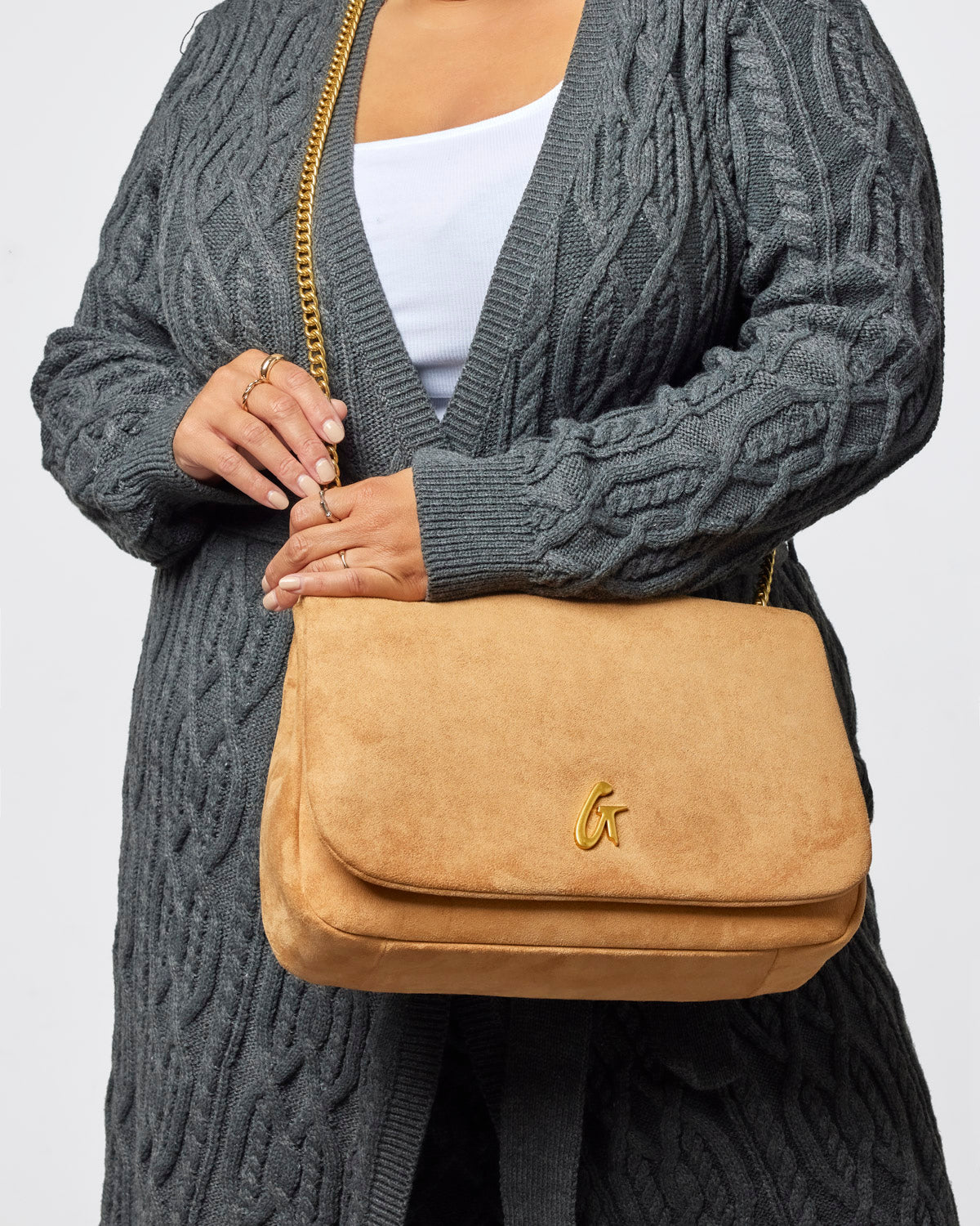 Medium tan suede flap bag with a gold logo, styled with a cozy gray cardigan for a chic look.