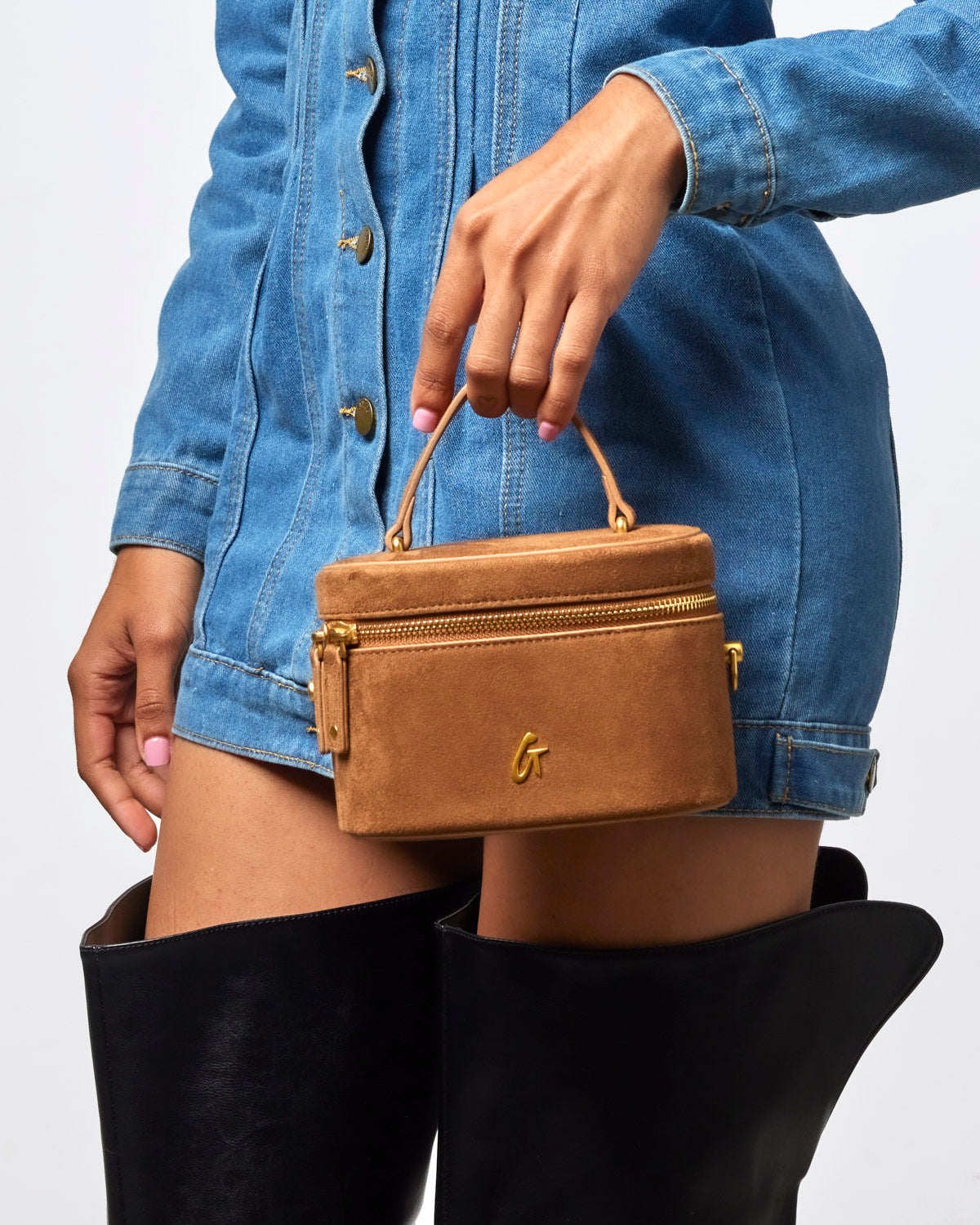 Brown mini suede vanity bag with gold zipper, held by a model wearing a denim dress and knee-high black boots.
