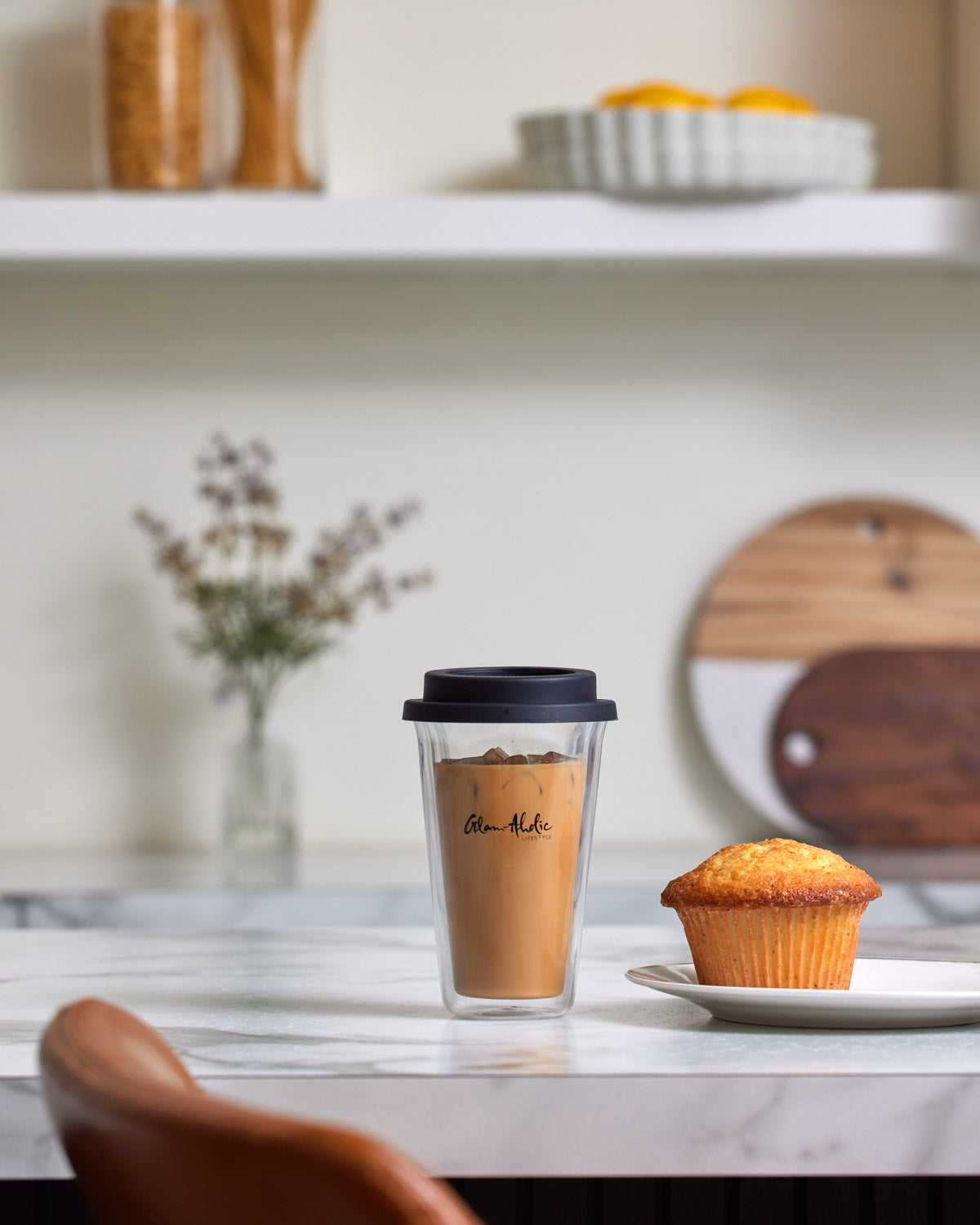 Clear signature logo tumbler with black lid, filled with iced coffee, on a white marble table next to a muffin.
