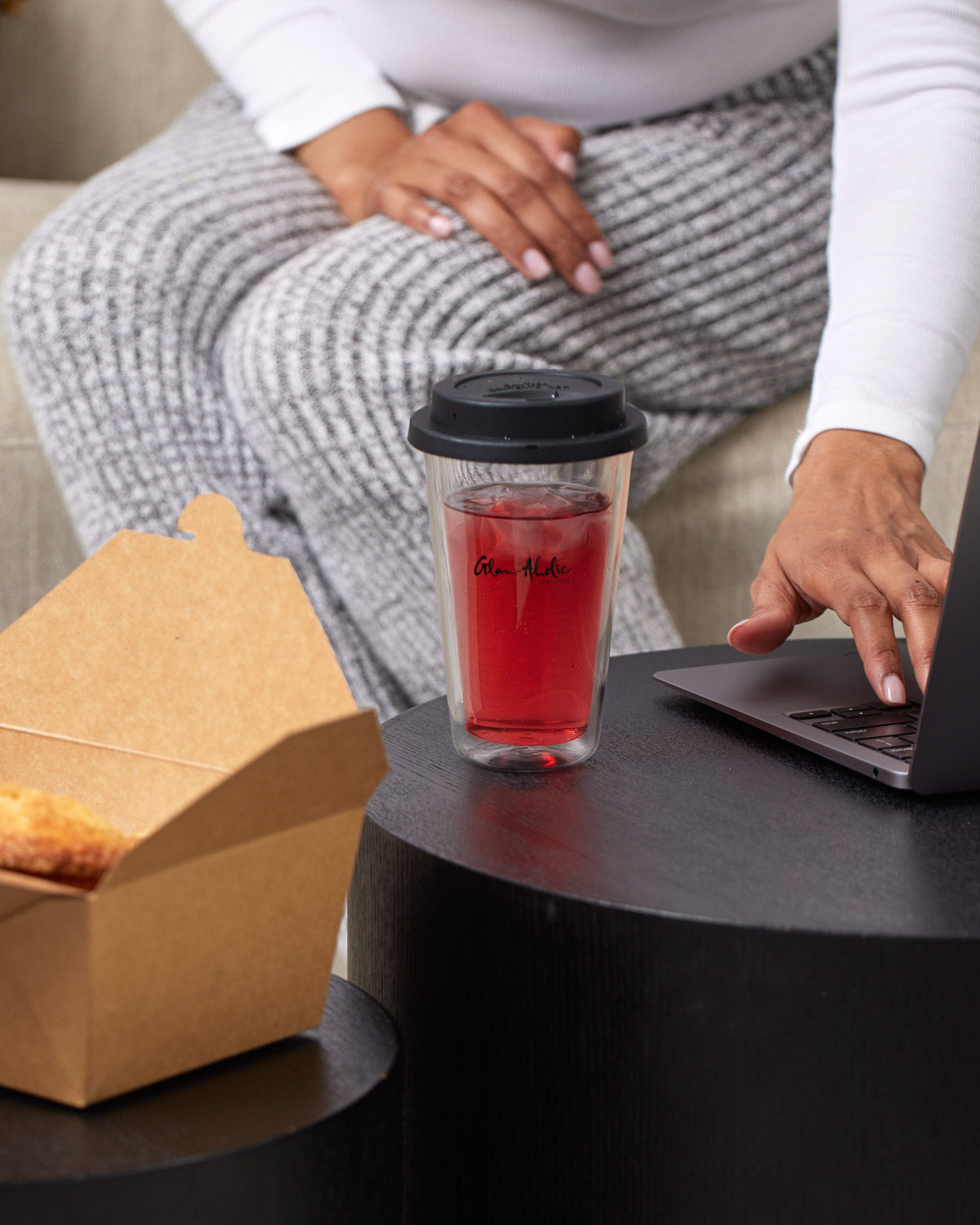 A clear tumbler with a black lid, featuring a signature logo, filled with red beverage and ice, placed on a black table.
