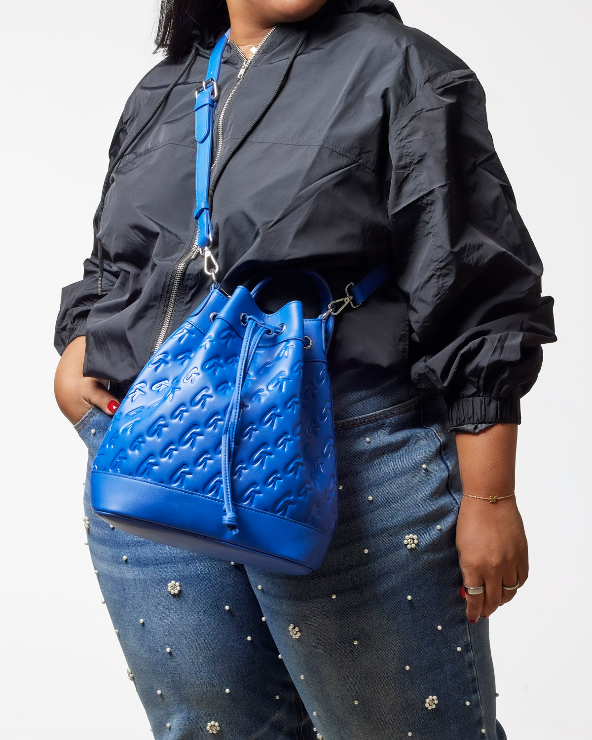 Large blue monogram bucket bag with adjustable strap, styled with a black jacket and embellished jeans.