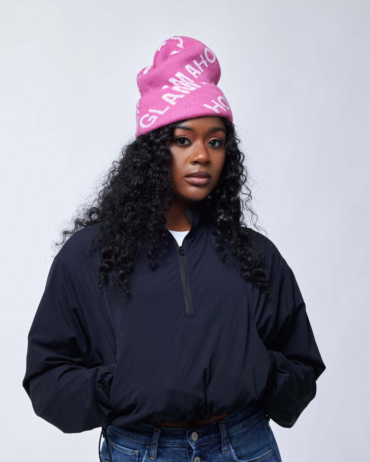 Woman wearing a black jacket and a bright pink Monogram Beanie with white lettering, styled casually against a plain background.