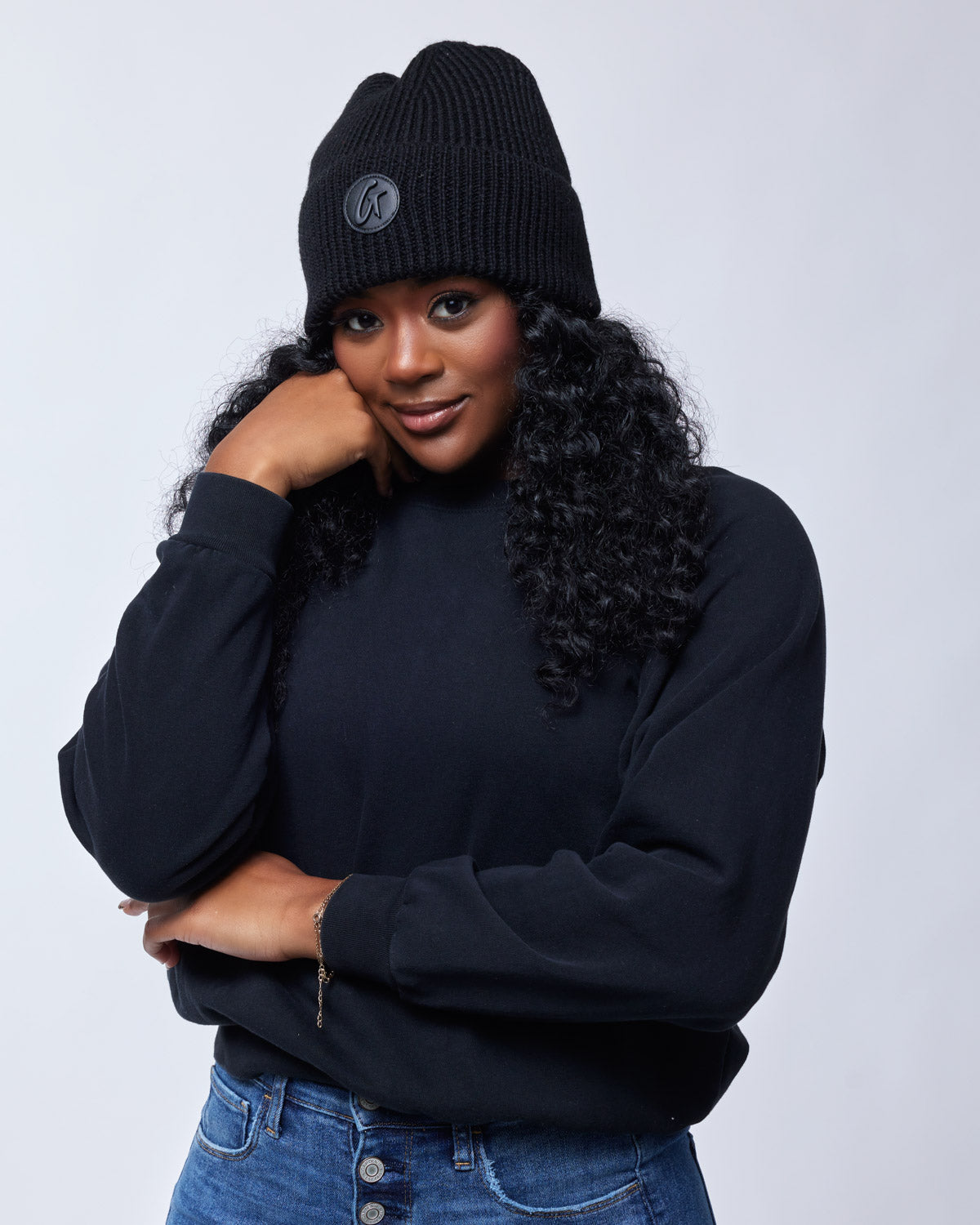 Woman wearing a black knit beanie with a logo, black sweatshirt, and blue jeans, posing with her hand near her face.