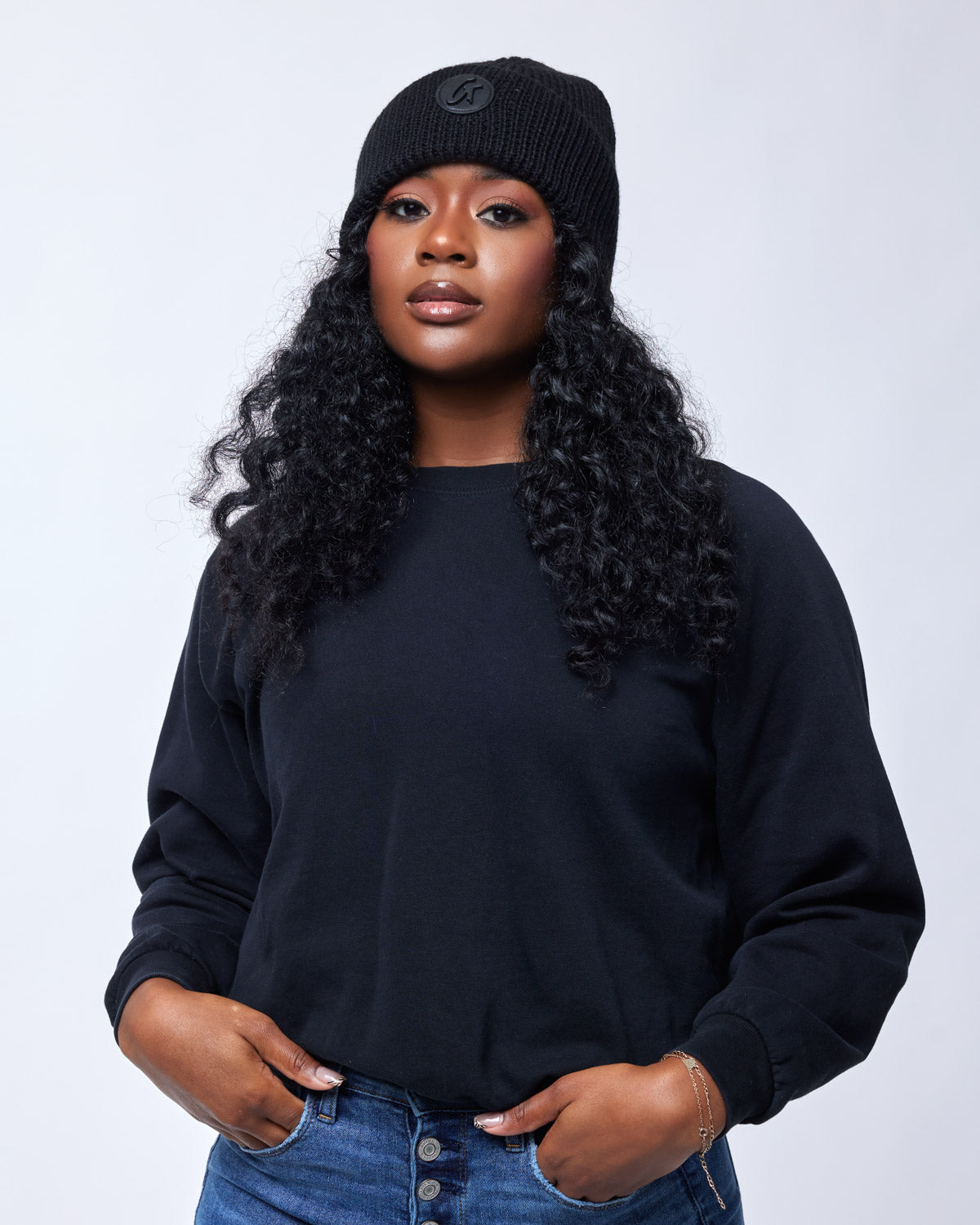 Black classic beanie hat with a logo patch, worn by a woman with curly hair, black sweatshirt, and blue jeans.