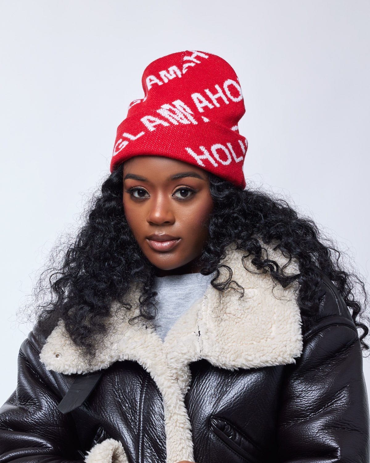Red monogram beanie with white lettering, worn by a woman with curly black hair, styled with a shearling-lined leather jacket.