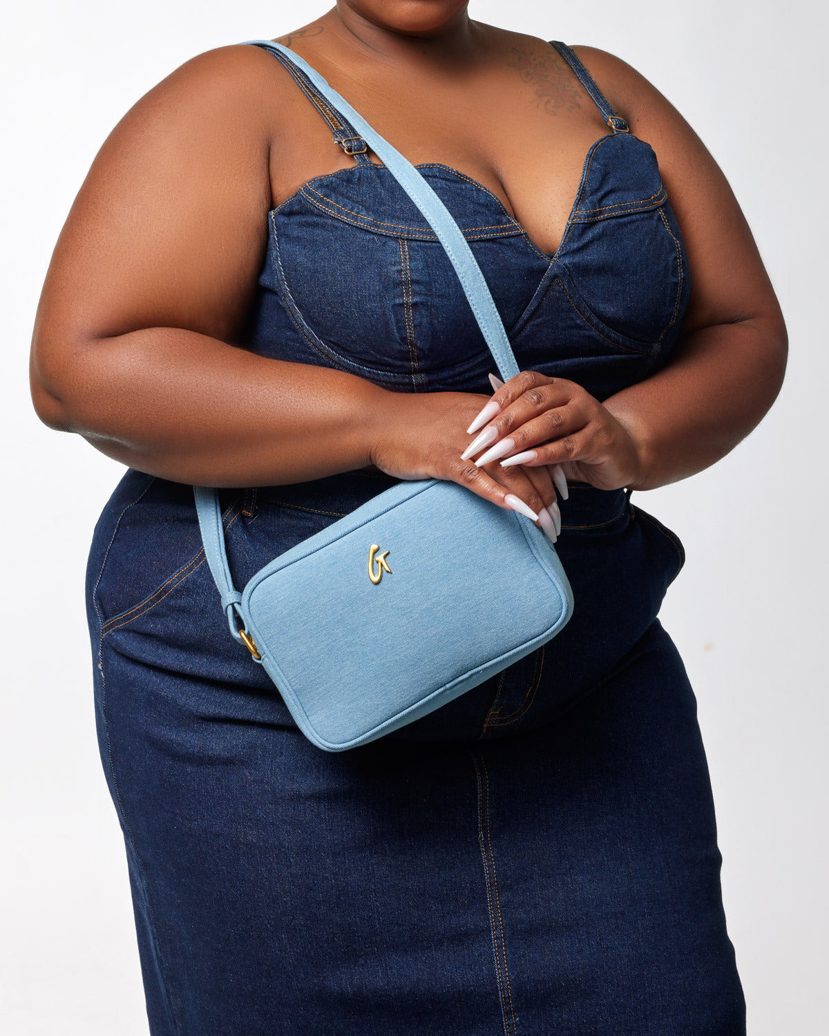 Light blue denim camera bag with a gold logo, worn crossbody by a woman in a denim outfit.