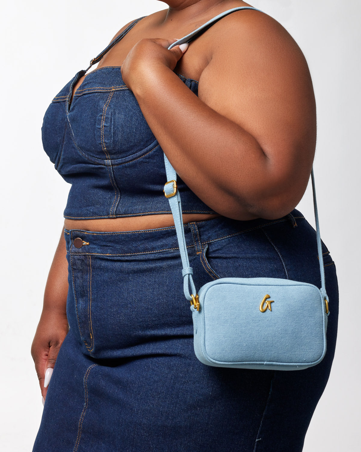 Mini denim gold camera bag with a light blue fabric exterior, gold logo, and adjustable strap, worn crossbody by a woman.