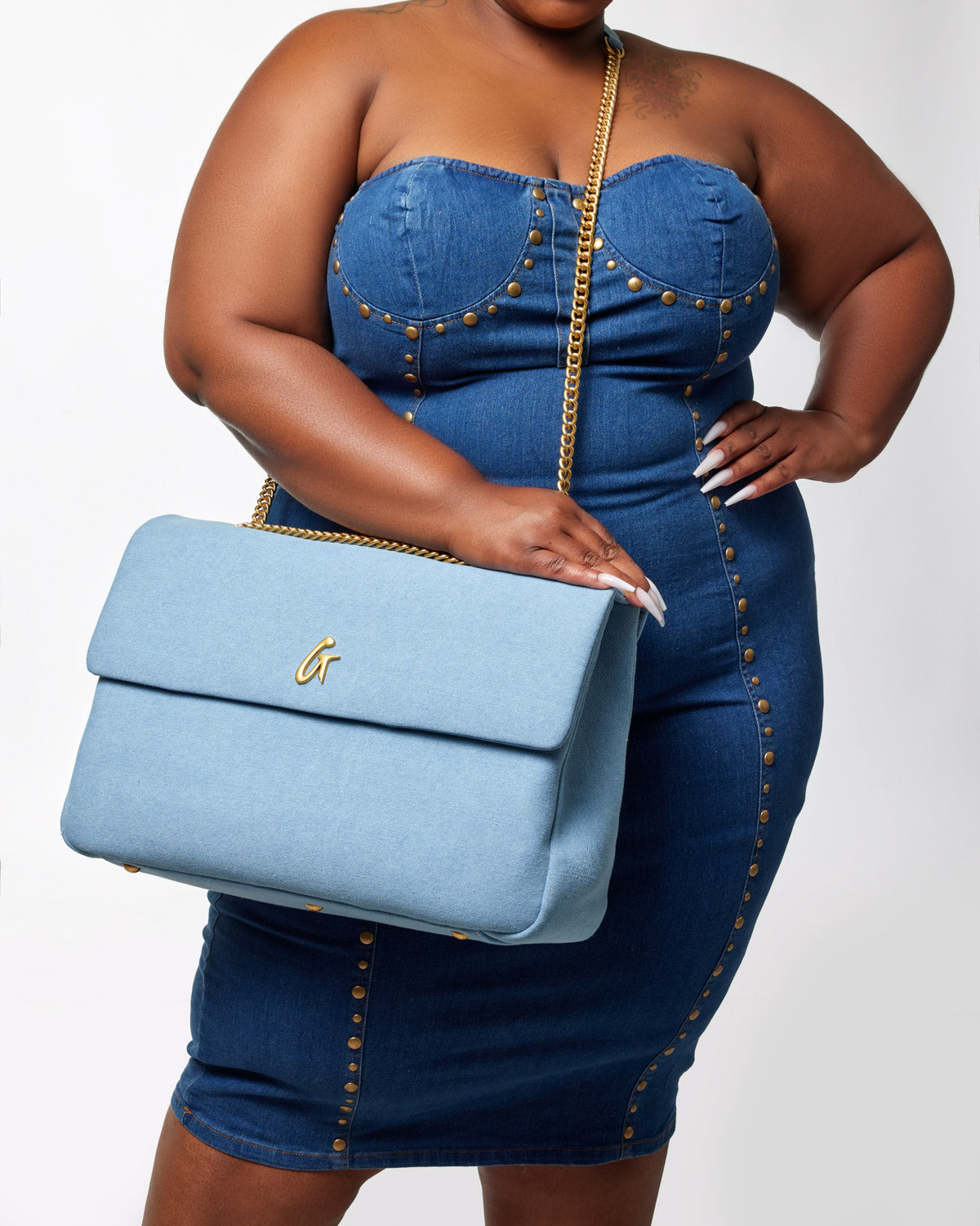 Large light blue denim flap bag with gold chain strap and gold logo detail, held by a woman wearing a denim dress with gold studs.