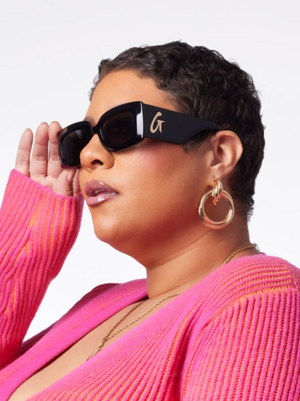 Stylish woman wearing black sunglasses with a gold logo, paired with a vibrant pink sweater and hoop earrings.