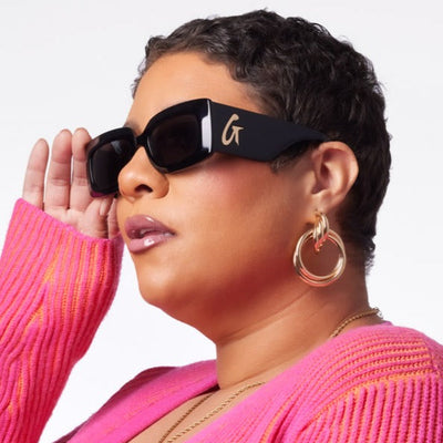 Stylish woman wearing black sunglasses with a gold logo, paired with a vibrant pink sweater and hoop earrings.