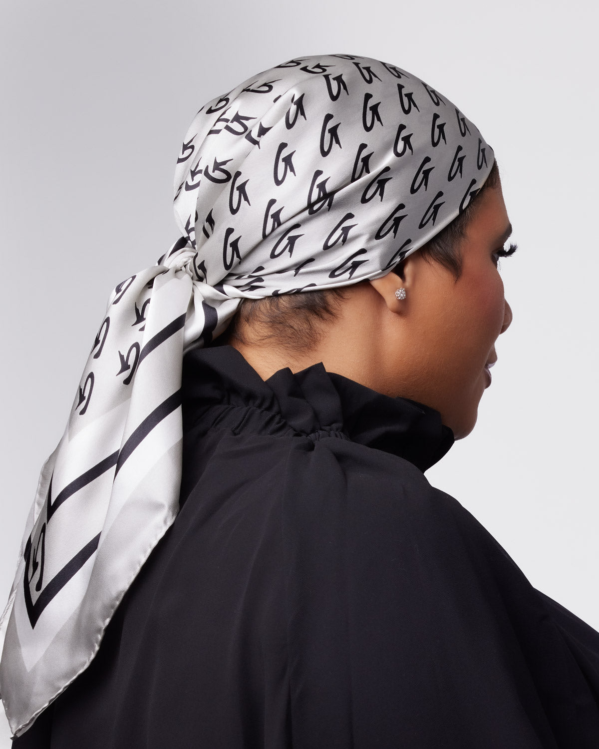 Large silver monogram silk scarf elegantly tied around a woman's head, featuring a stylish pattern of black letters.