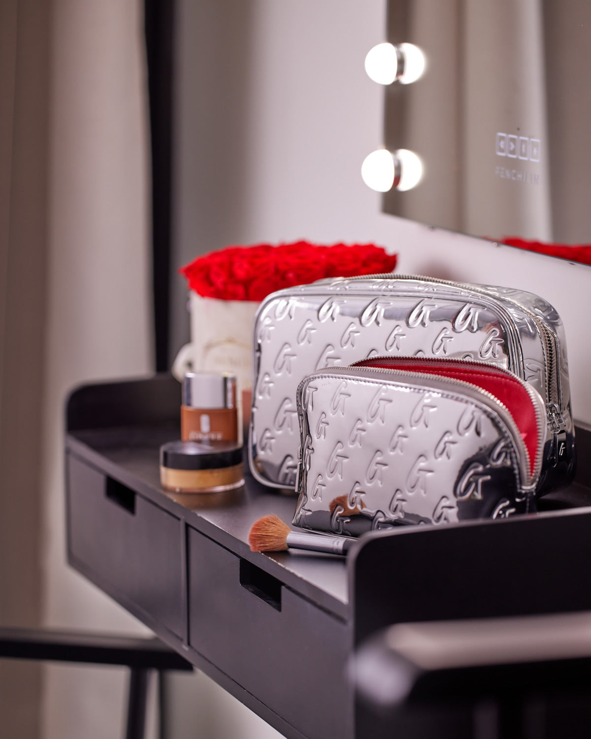 Small metallic silver monogram toiletry bag with embossed G pattern, placed on a black vanity table with makeup items.