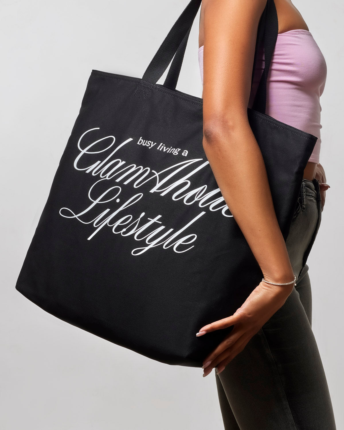 Black canvas tote bag featuring the phrase "busy living a Glamhouse Lifestyle," held by a woman.