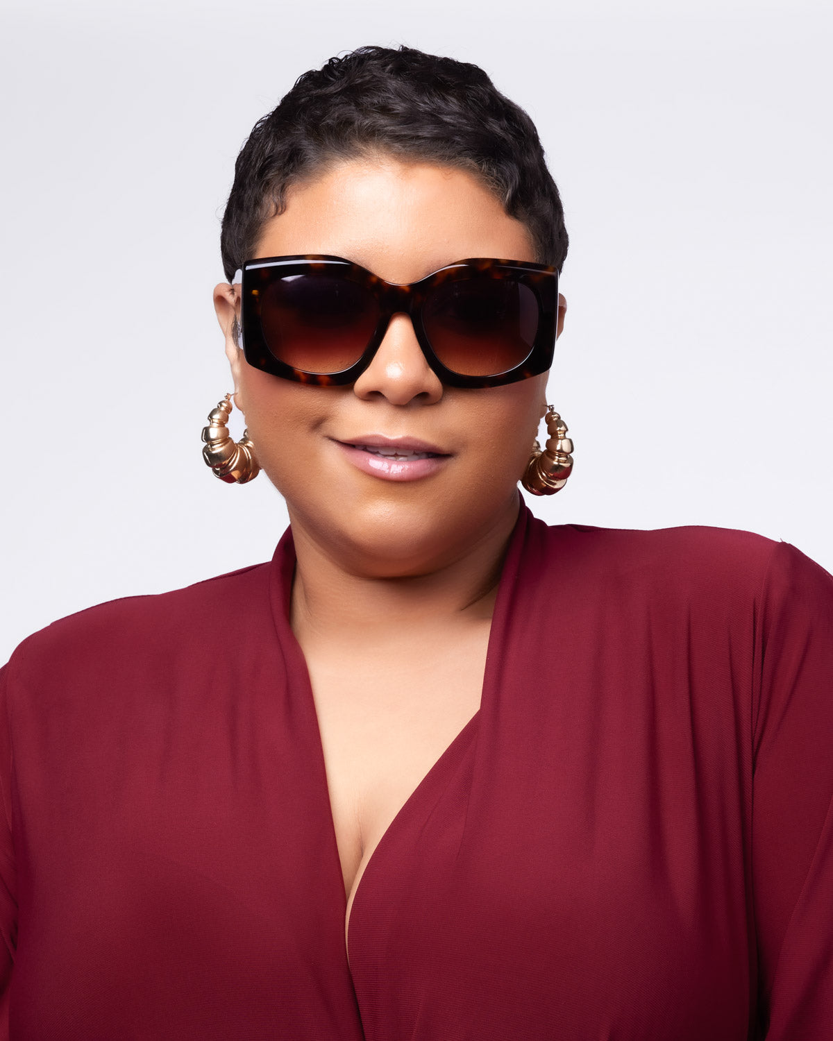 Model wearing oversized tortoise sunglasses and statement gold earrings, styled in a deep burgundy outfit.