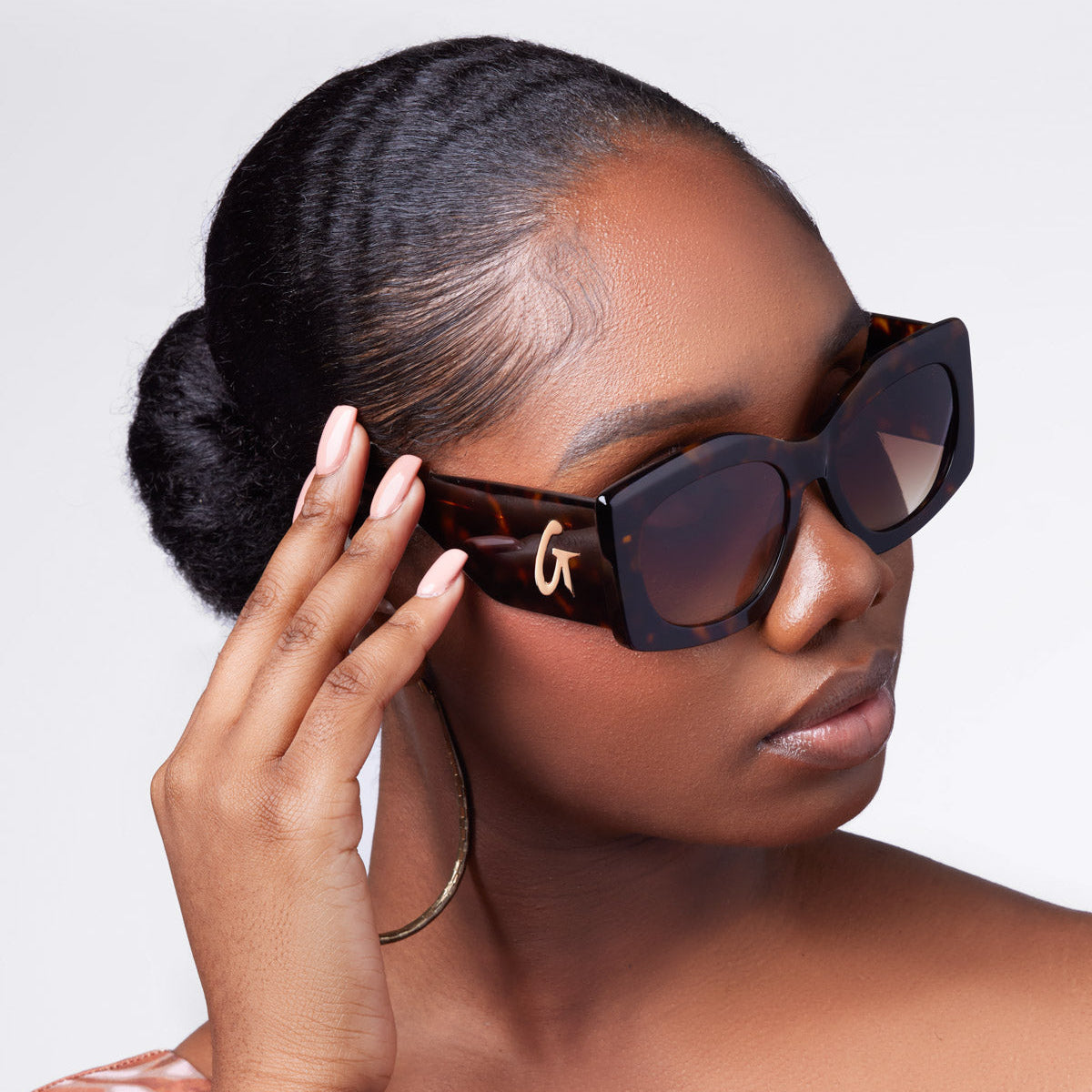 A woman wearing oversized tortoise sunglasses, gently touching the frame, with a stylish off-shoulder top and elegant hairstyle.