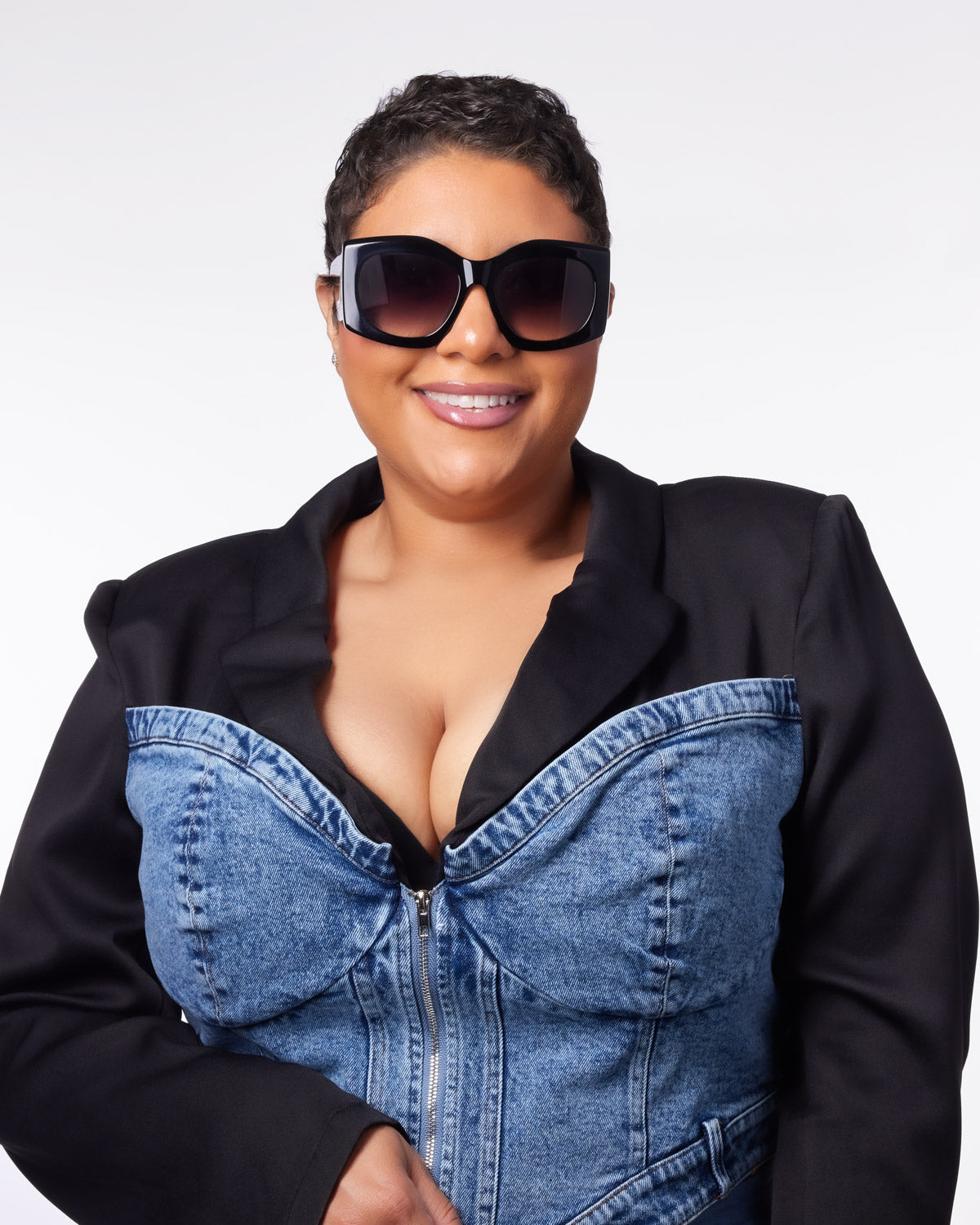 Stylish woman wearing oversized black sunglasses and a denim corset jacket, smiling confidently against a white background.