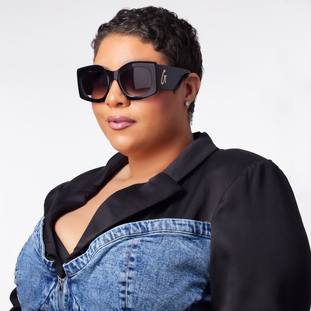 Stylish black oversized sunglasses with a gold logo, worn by a woman with short hair and a chic outfit.