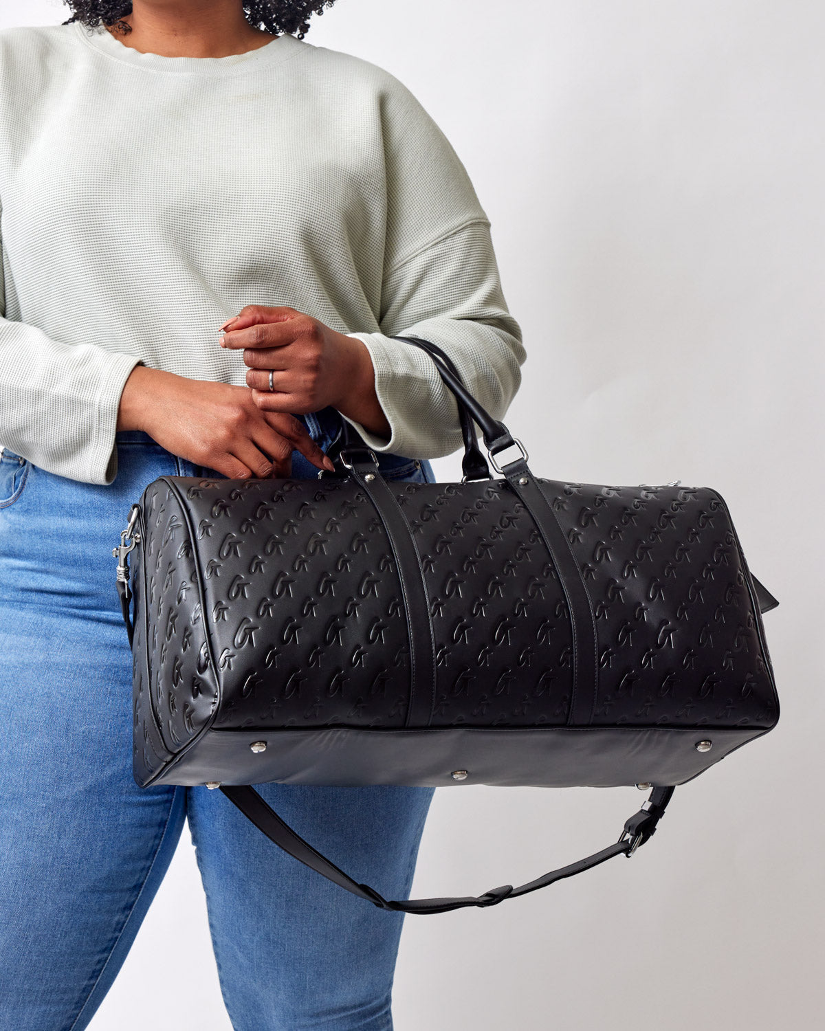 Black monogram unisex duffle bag with embossed logo pattern, carried by a person in casual clothing.