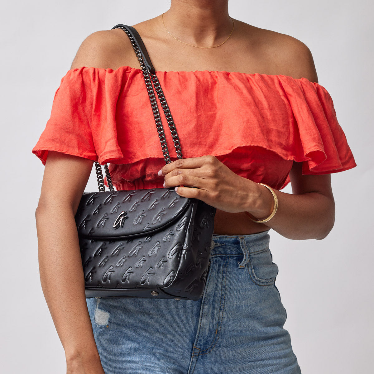 Black medium monogram flap bag with embossed logo pattern, worn over a red off-shoulder top and blue jeans.