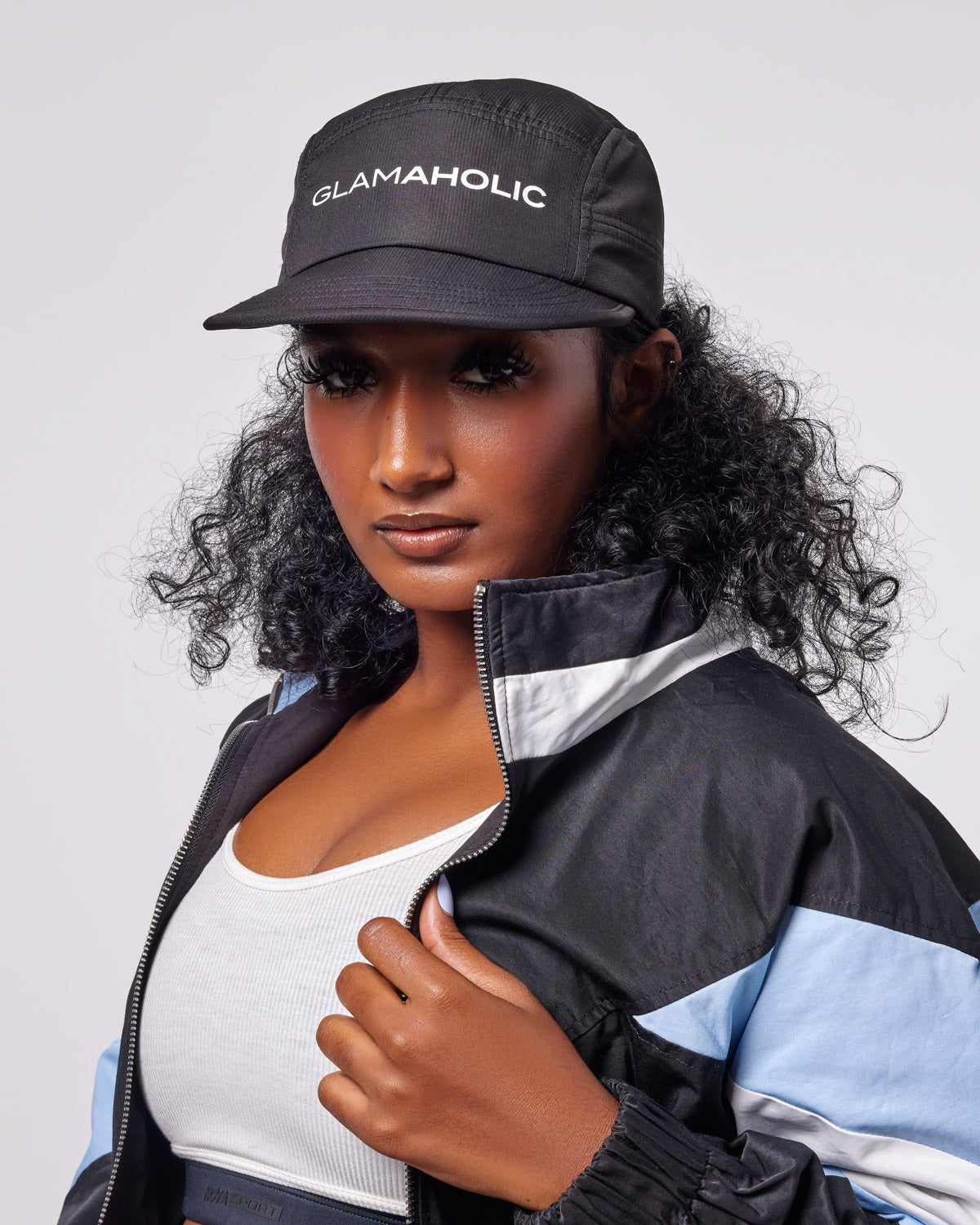 Model wearing a black athletic 5 panel hat with "GLAMAHOLIC" logo, styled with a sporty jacket and curly hair.