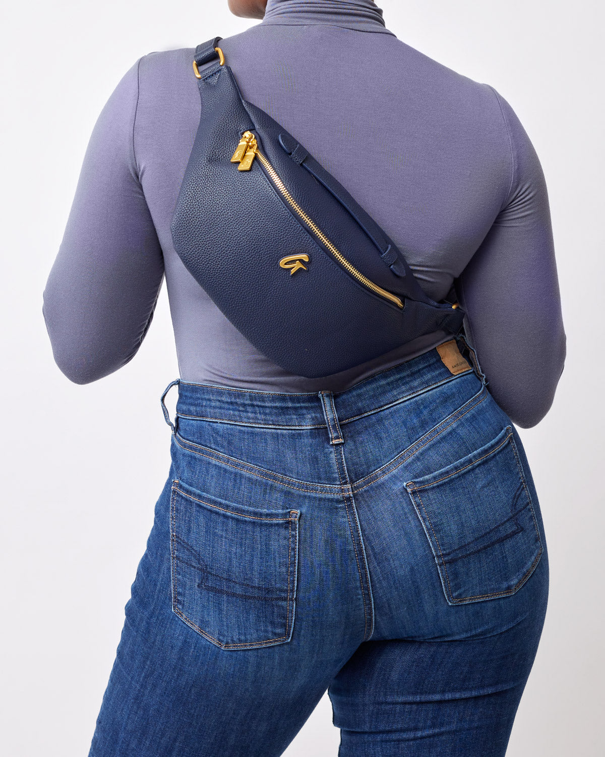 Navy Pebble Fanny Pack worn across the chest, featuring gold zippers and logo, styled with blue jeans and a gray top.
