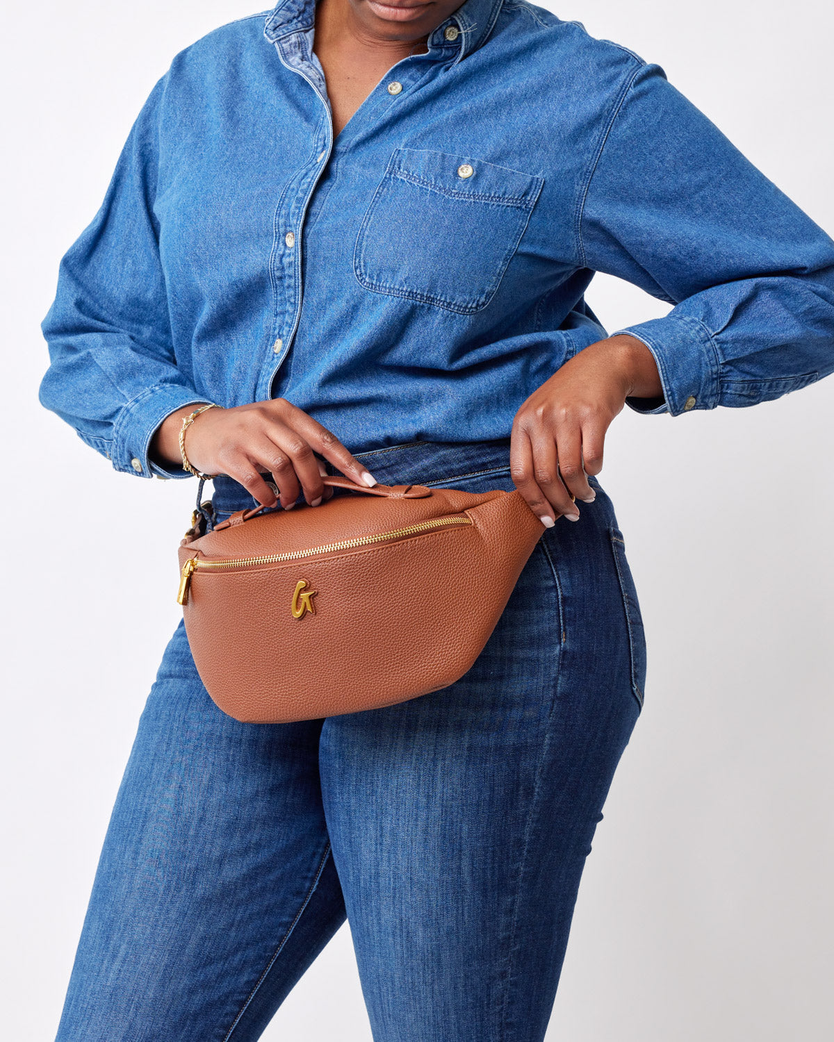 Brown pebble-textured fanny pack with gold zipper and logo, worn around the waist of a person in a denim shirt and jeans.