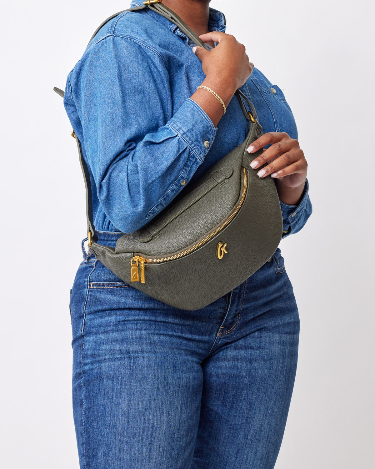 Olive green pebble-finish fanny pack with gold zipper and logo, worn across the body over a denim jacket and jeans.