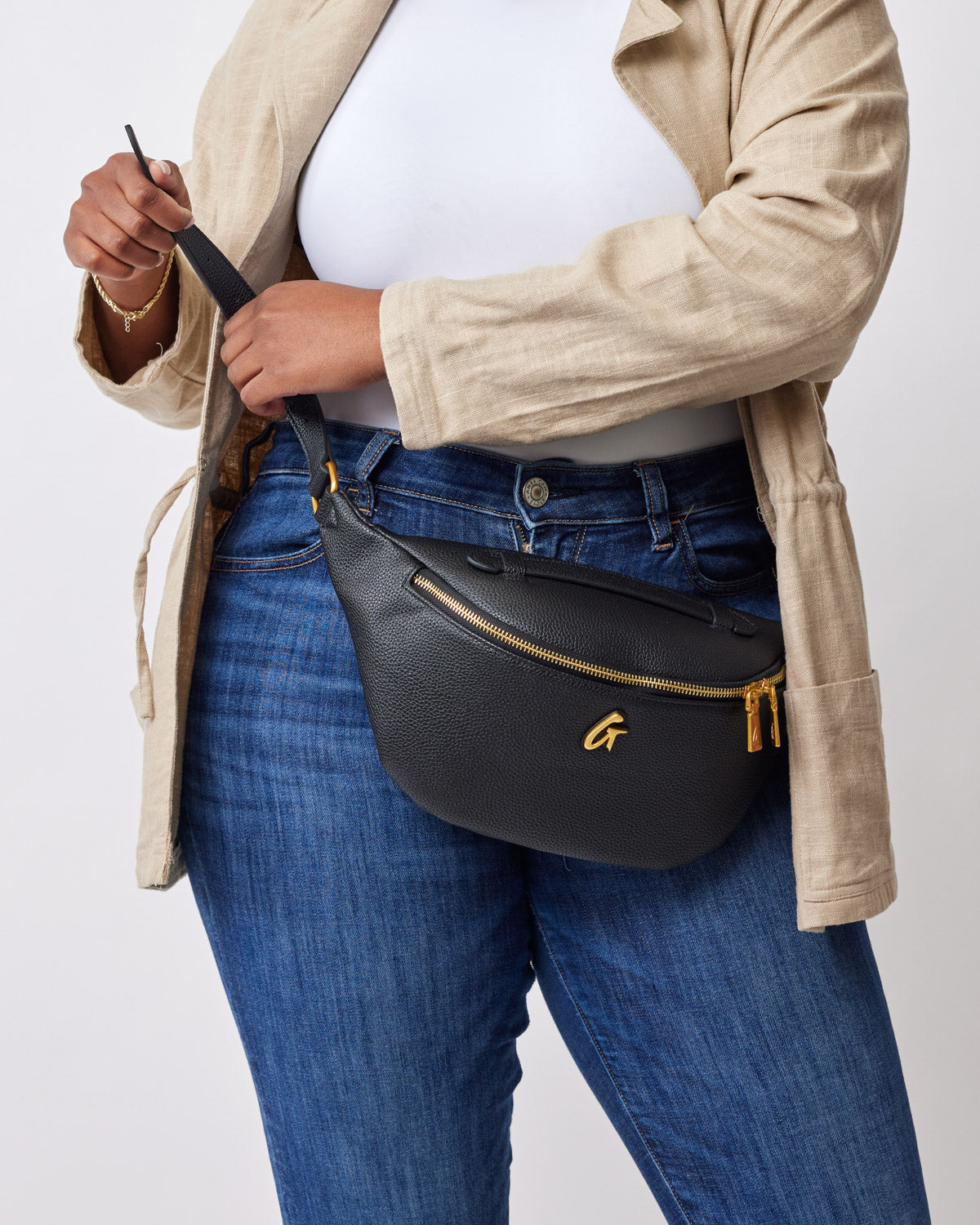 Black Pebble Fanny Pack with gold zipper and logo, worn around the waist over blue jeans and a beige jacket.