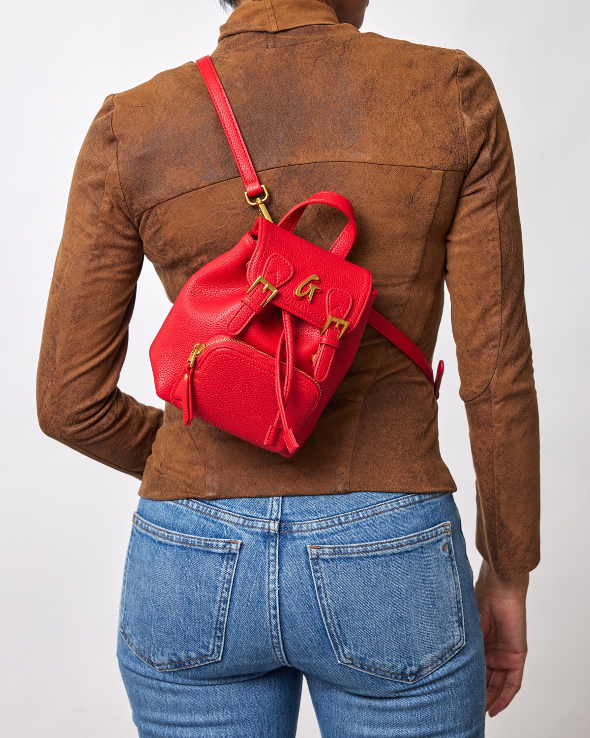 Red mini pebble crossbody backpack with gold hardware, adjustable strap, front zip pocket, and top handle, worn over a brown jacket.