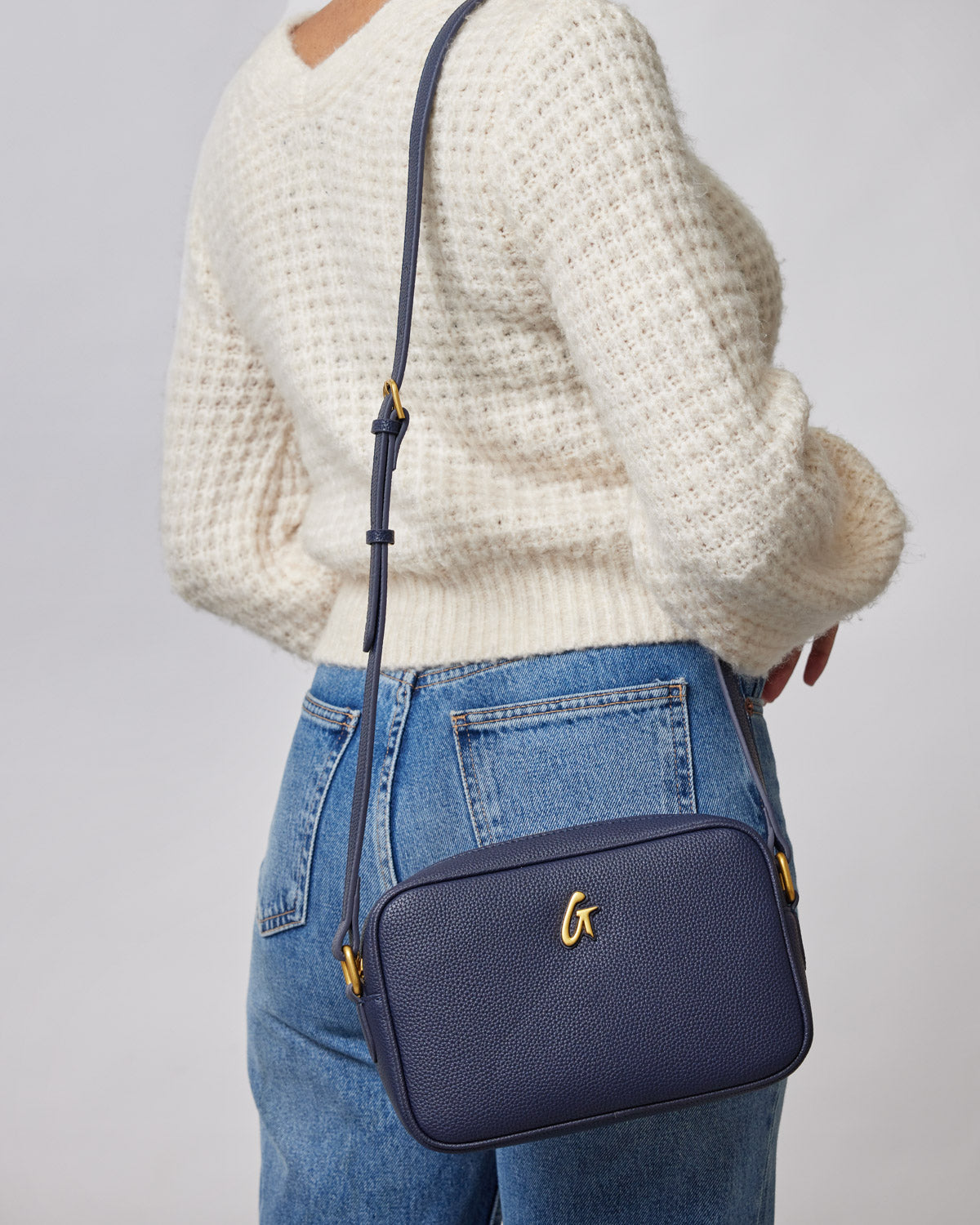 Navy pebble camera bag with adjustable strap worn crossbody over a cream sweater and blue jeans.