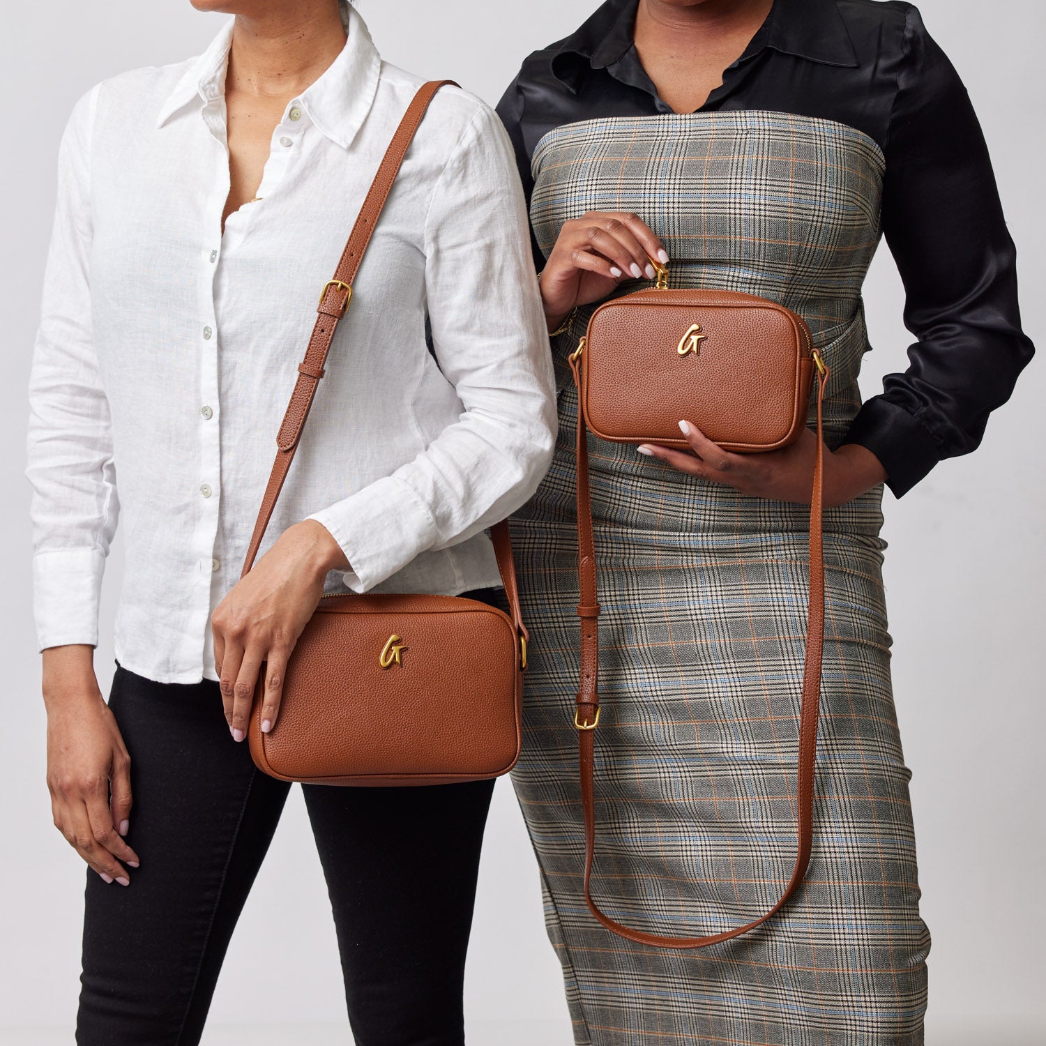 Two women hold brown mini pebble camera bags with gold logo and adjustable straps, showcasing stylish accessories.