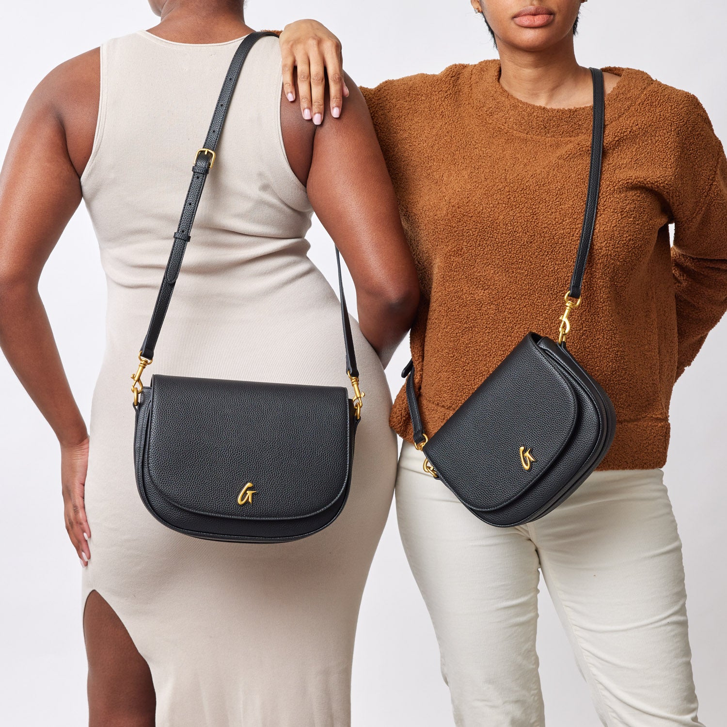 Two women display black pebble leather shoulder bags with gold hardware and adjustable straps, styled with casual outfits.