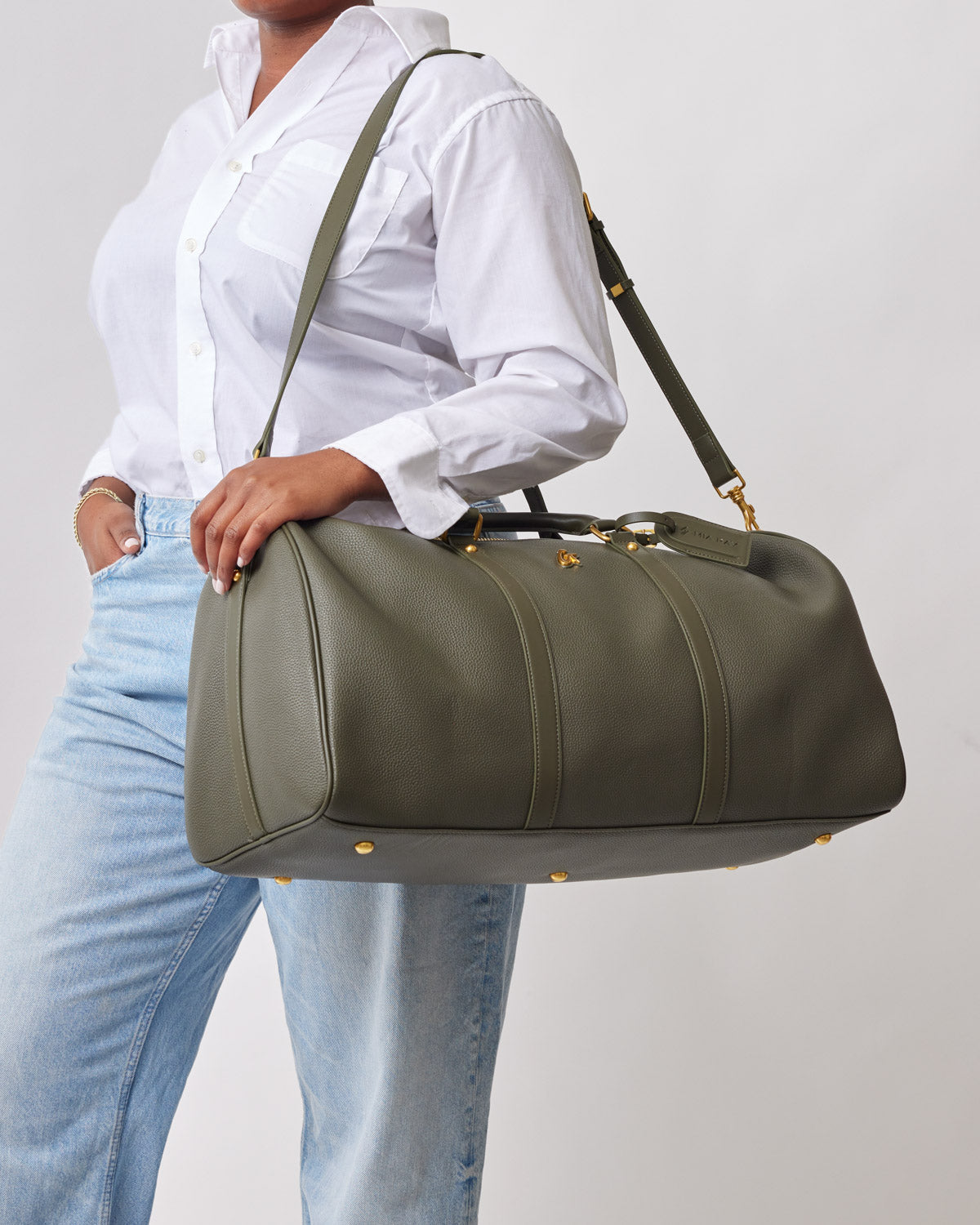 Olive green Pebble Duffle Bag with gold accents, carried by a person in a white shirt and light jeans.
