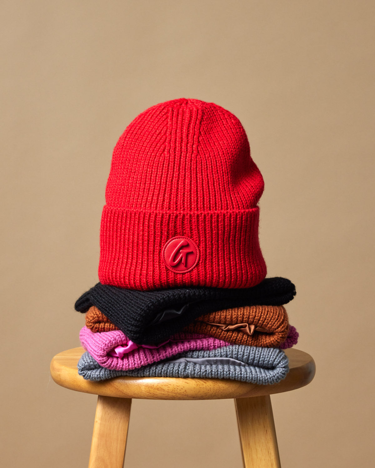 Red classic beanie displayed on top of a stack of colorful knit hats on a wooden stool.