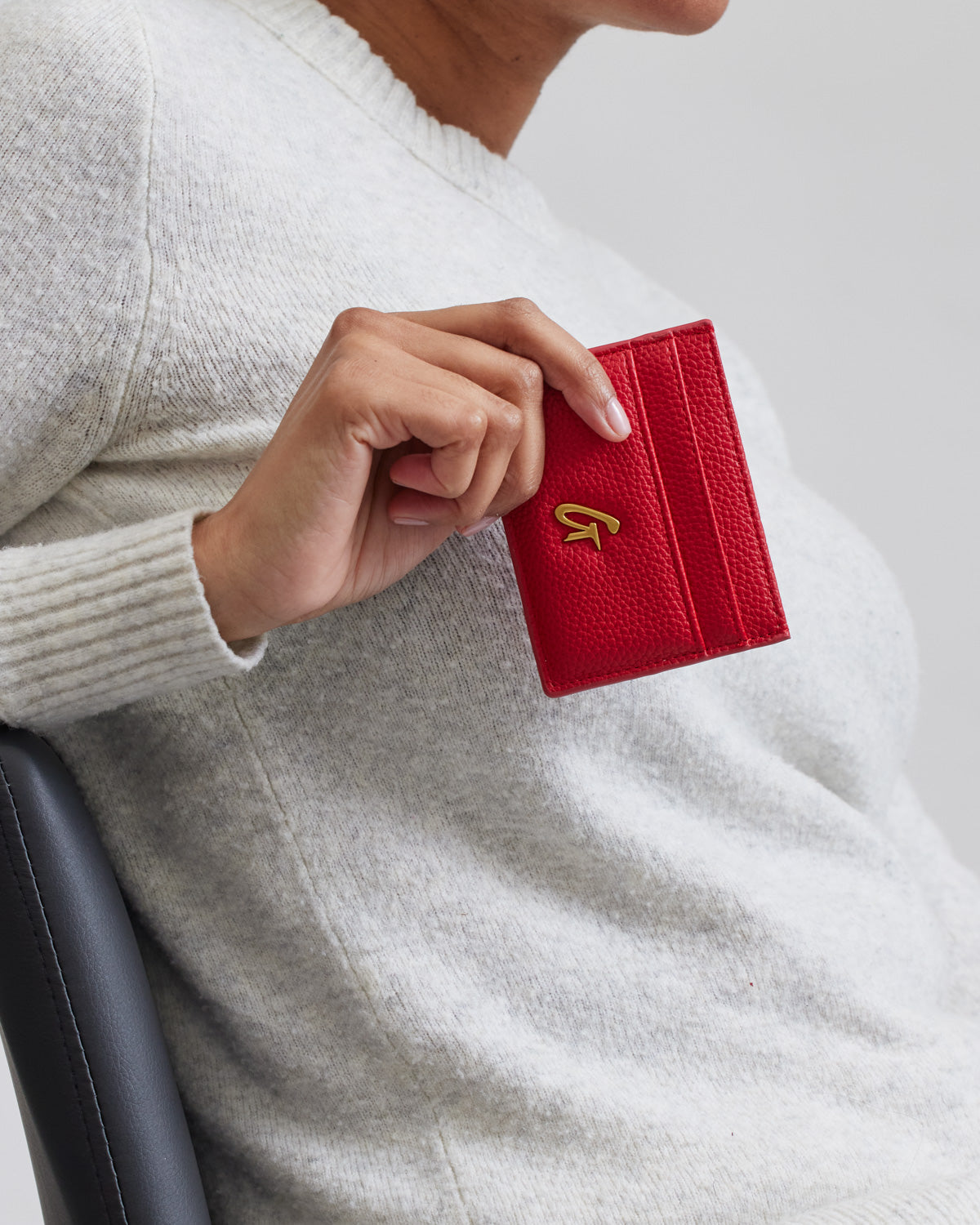 Red pebble leather card holder with a gold logo, held by a person wearing a cream sweater, close-up shot.