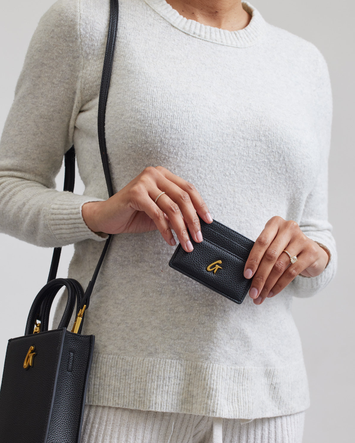 Black Pebble Card Holder with gold logo, held by a woman in a cream sweater, with a matching black bag.