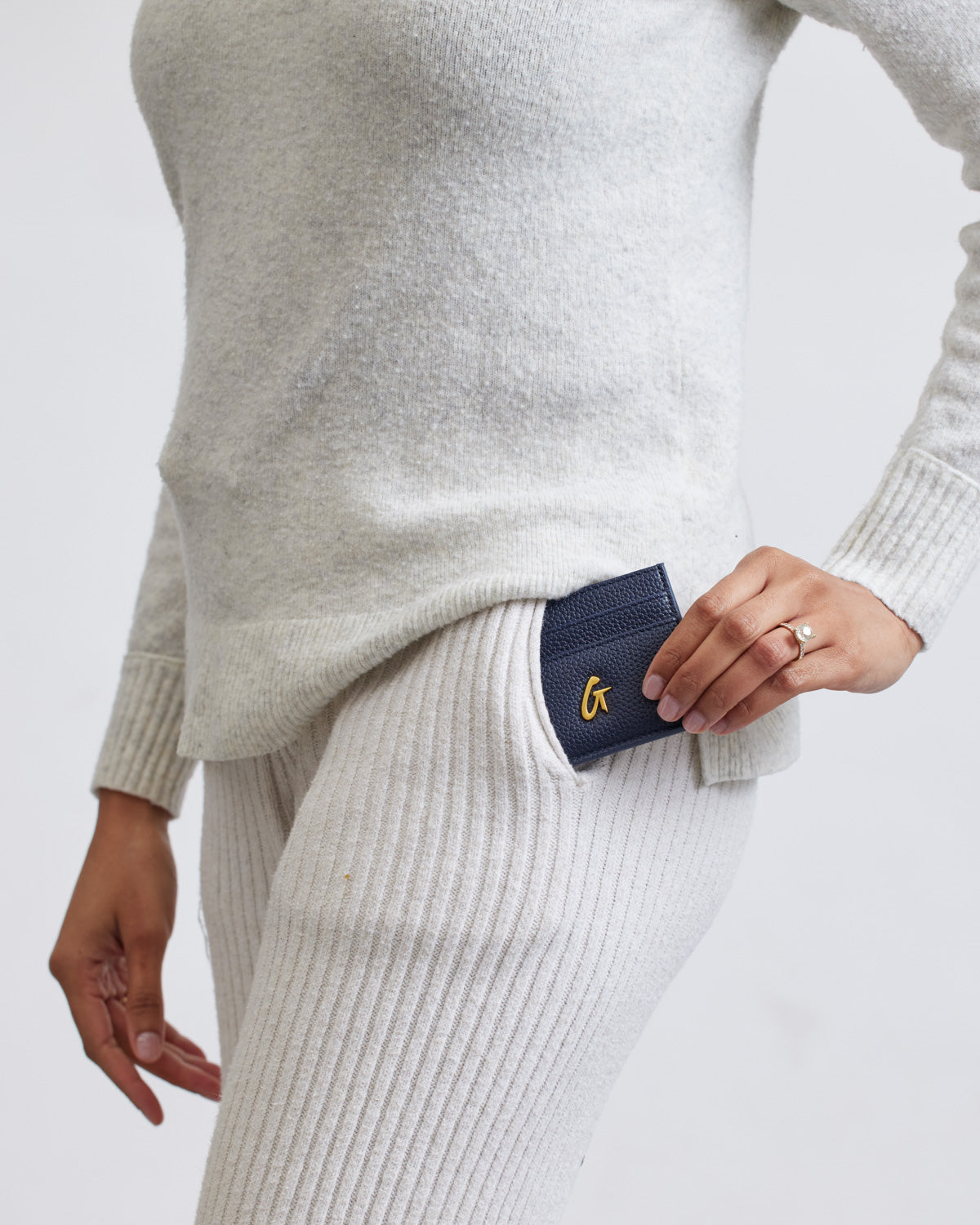Navy Pebble card holder partially tucked into the pocket of cream ribbed pants, with a gold logo and a ring on the person's finger.