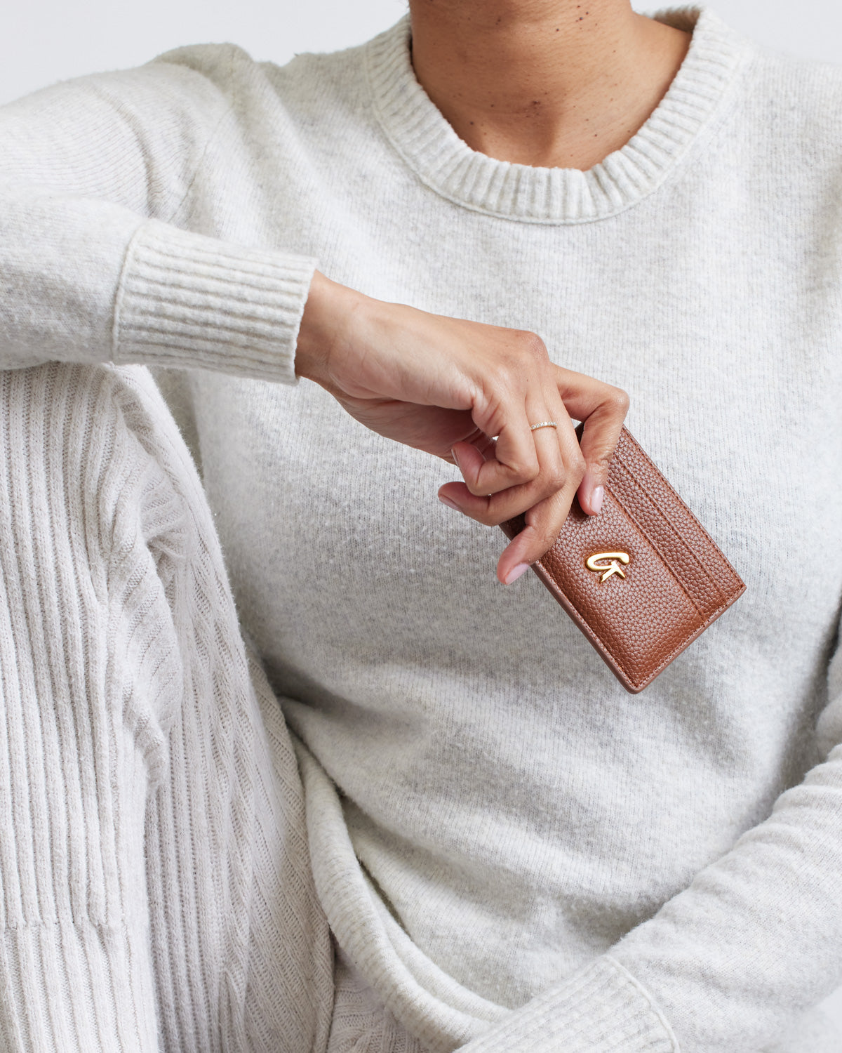 Close-up of a person holding a brown pebble card holder with a gold logo, wearing a cream sweater.