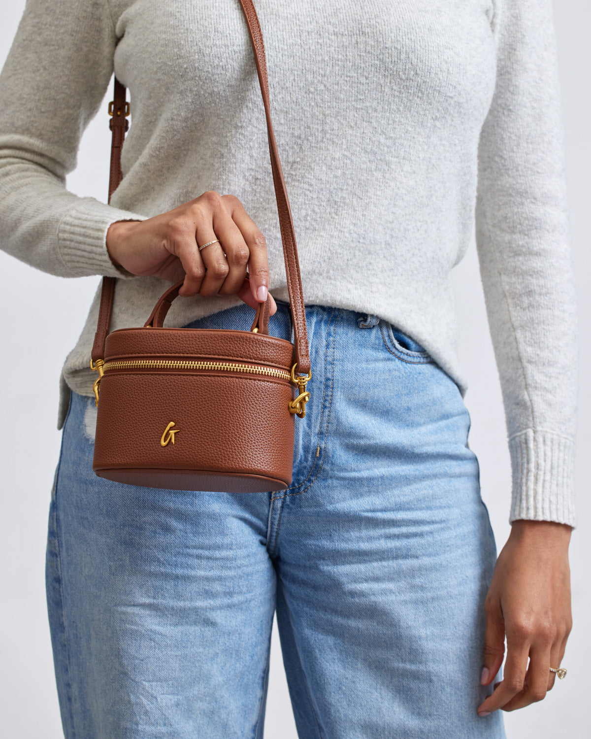 Mini pebble brown vanity bag with gold zipper and logo, held by a person wearing a gray sweater and blue jeans.