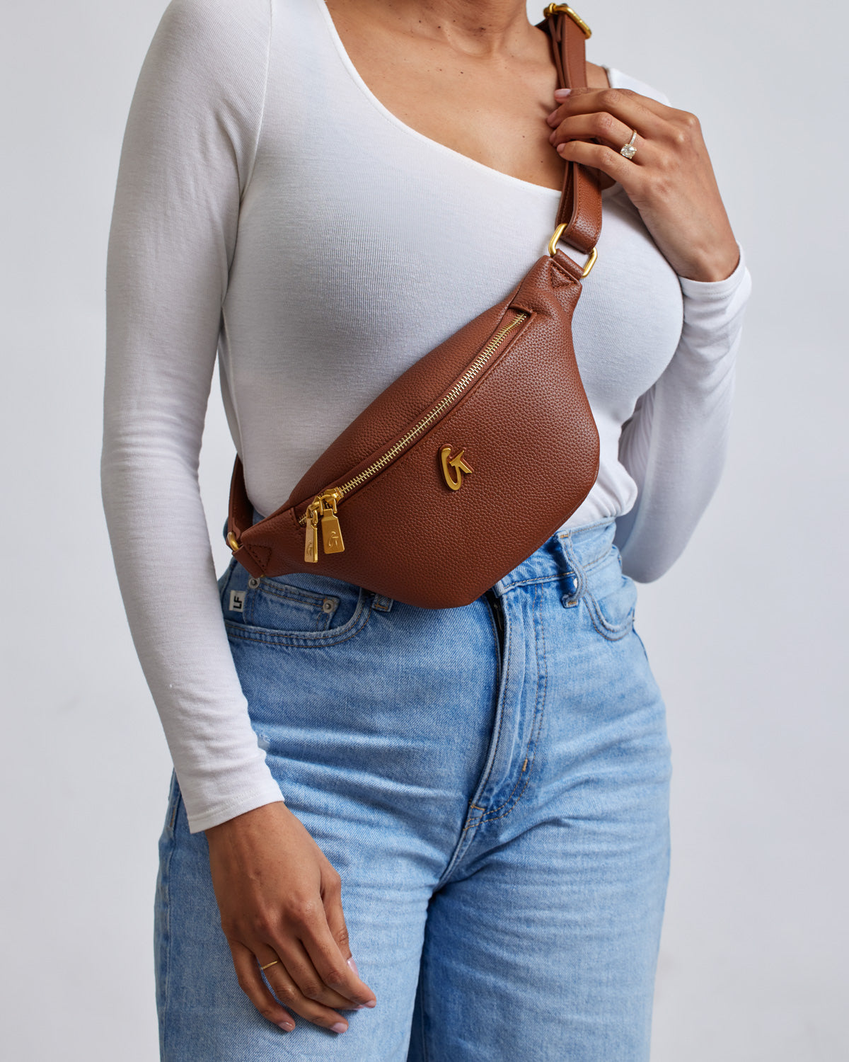Brown Pebble Fanny Pack bundle with gold zipper and logo, worn across the chest over a white long-sleeve top and blue jeans.