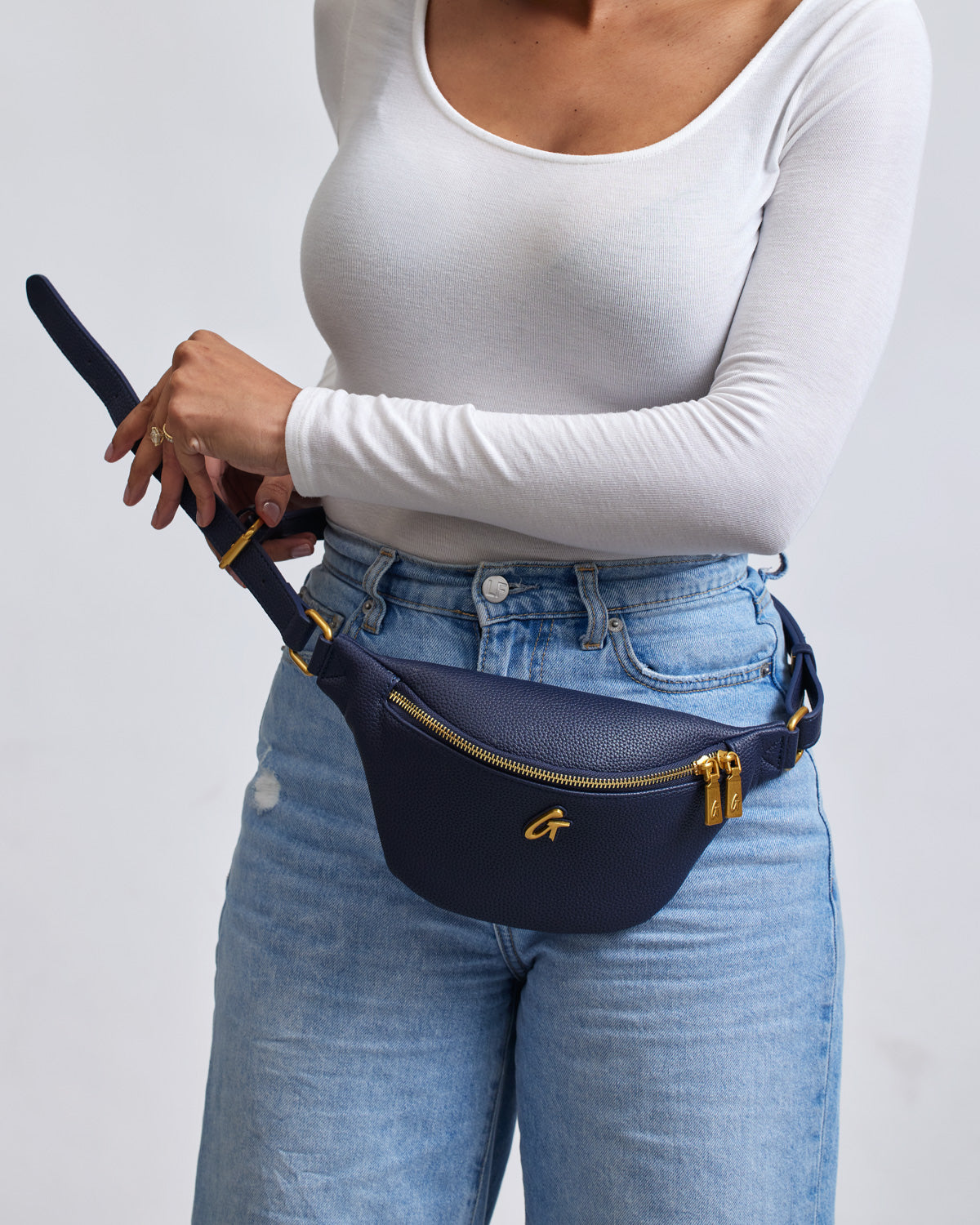 Navy mini pebble fanny pack with gold zipper and logo, worn around the waist of a person in a white long-sleeve top and jeans.