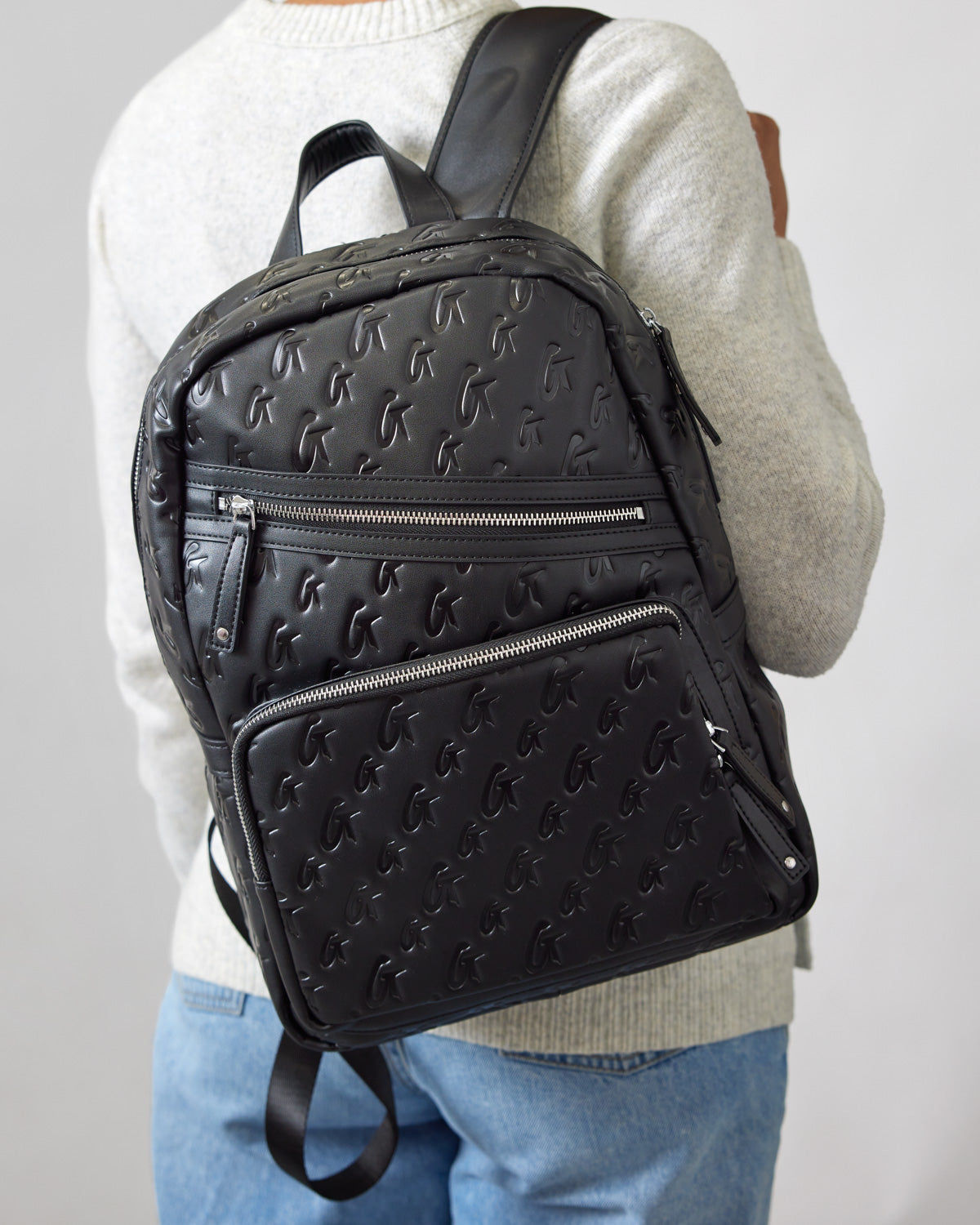 Black unisex backpack with embossed monogram pattern, multiple zippered compartments, and adjustable straps.