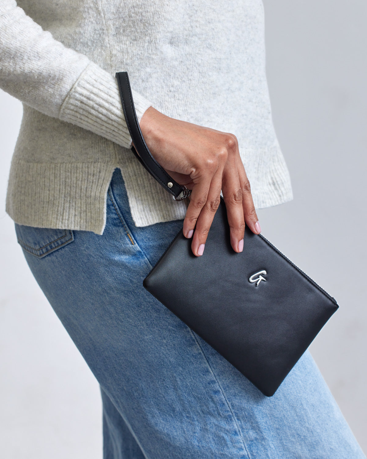 A person holds a black mini wristlet with a silver logo, wearing a beige sweater and blue jeans, against a plain background.